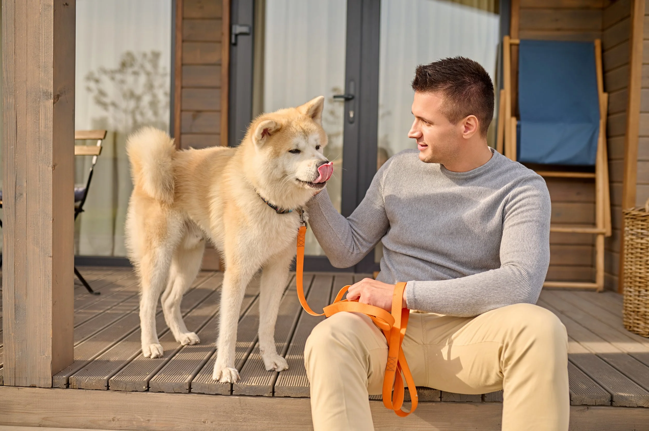 A man crouched on a wooden deck petting a fluffy cream-colored dog with a black collar and orange leash, near a wooden post and a door, with a blue shade in the background.