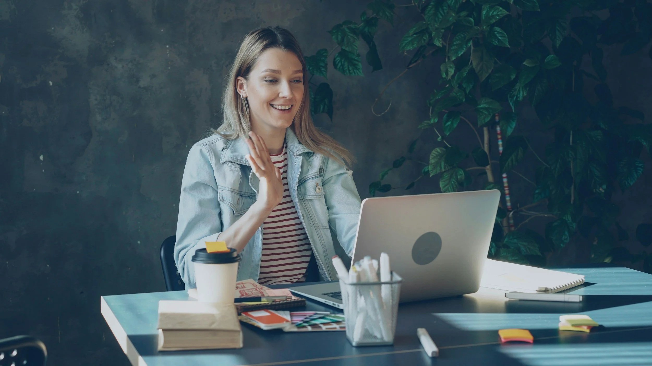 A young woman with blonde hair smiling and waving while sitting at a desk with a laptop. The desk has various office supplies, a coffee cup, and a notebook. There are plants and a textured wall in the background.