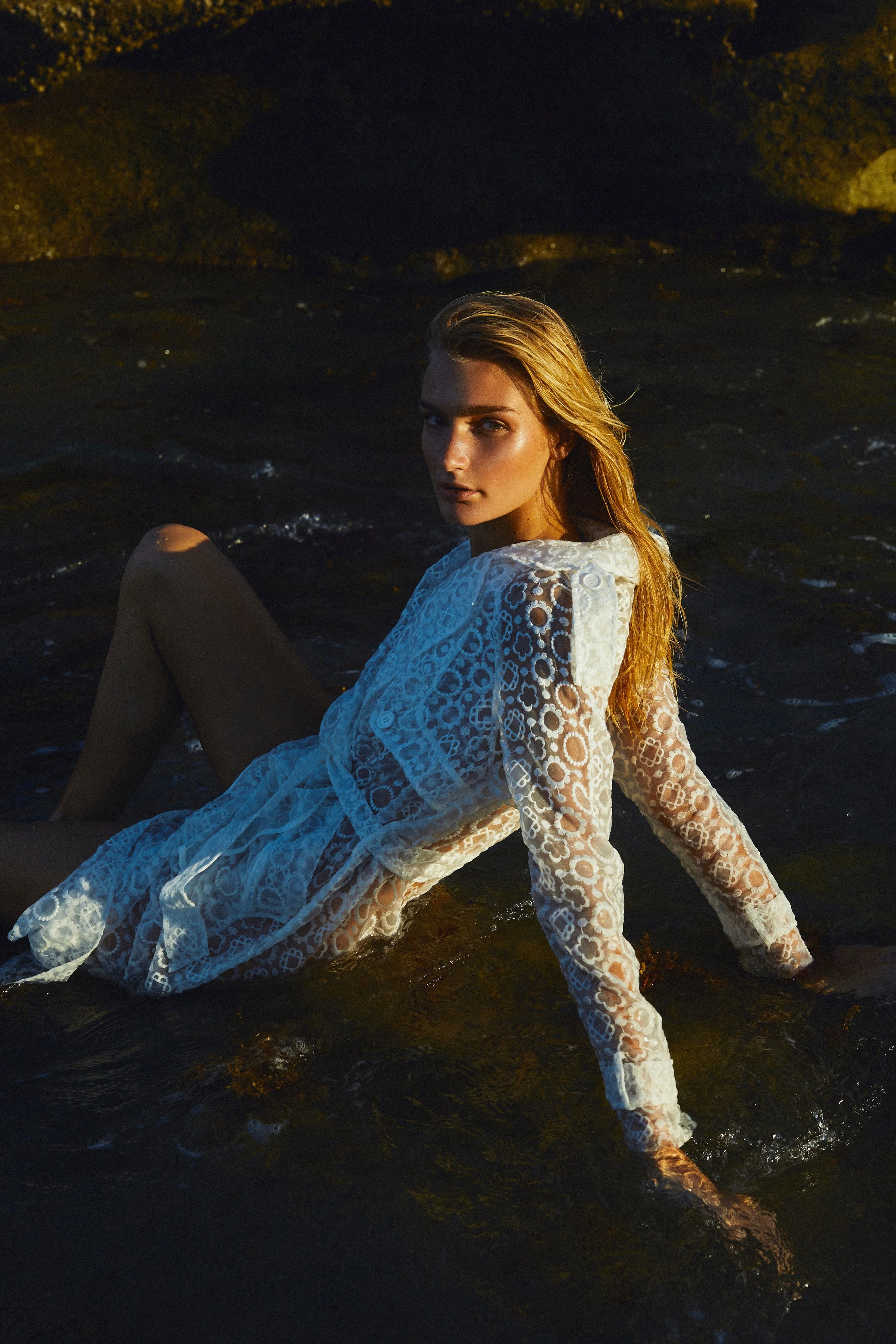 A young woman with blonde hair sitting in shallow water at sunset, wearing a sheer, lace, white dress.