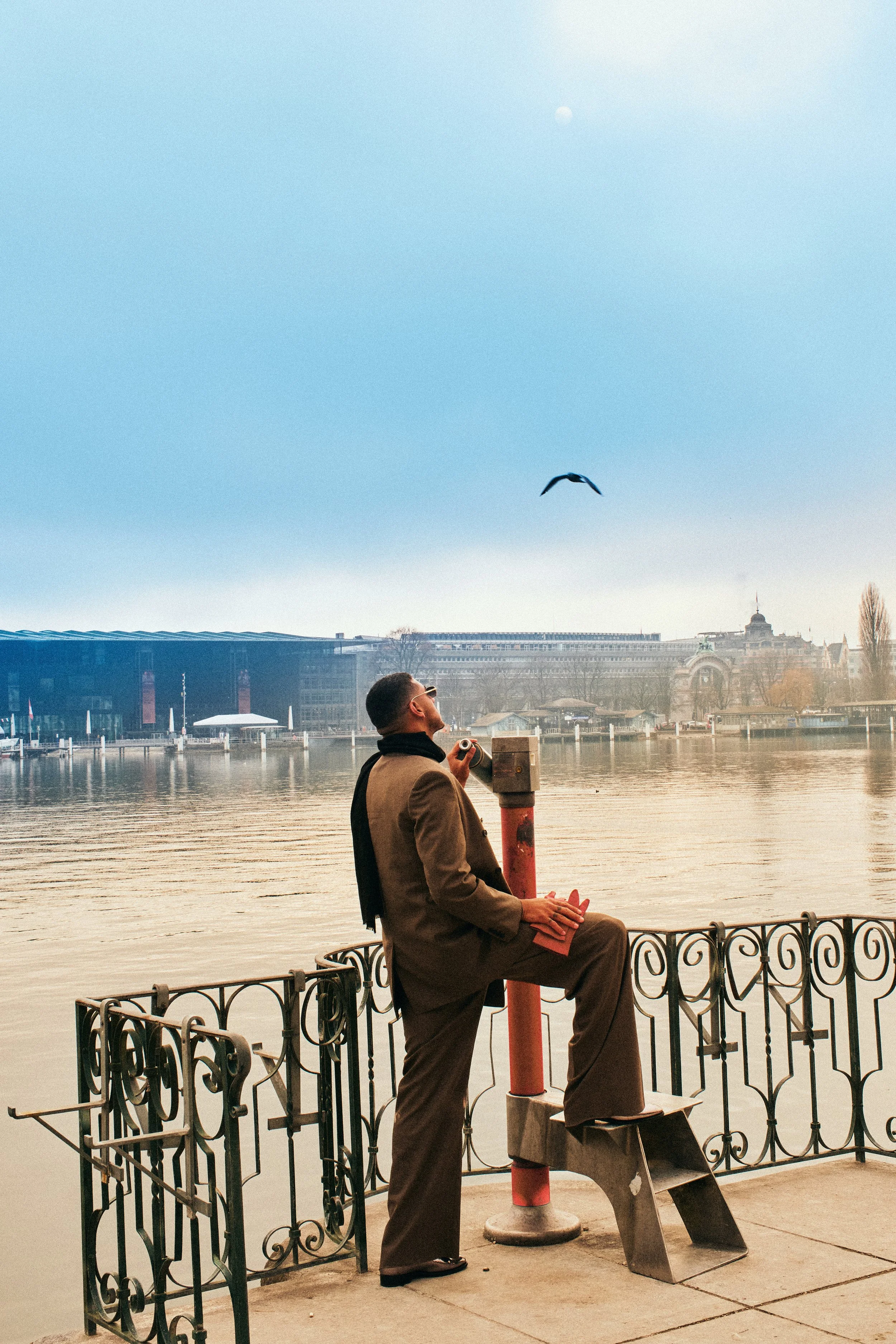 A man in a brown suit and black scarf stands by a river, holding a camera, with one foot resting on a small wooden step stool. He is looking up at the sky where a bird is flying, and the moon is visible in the clear daytime sky.