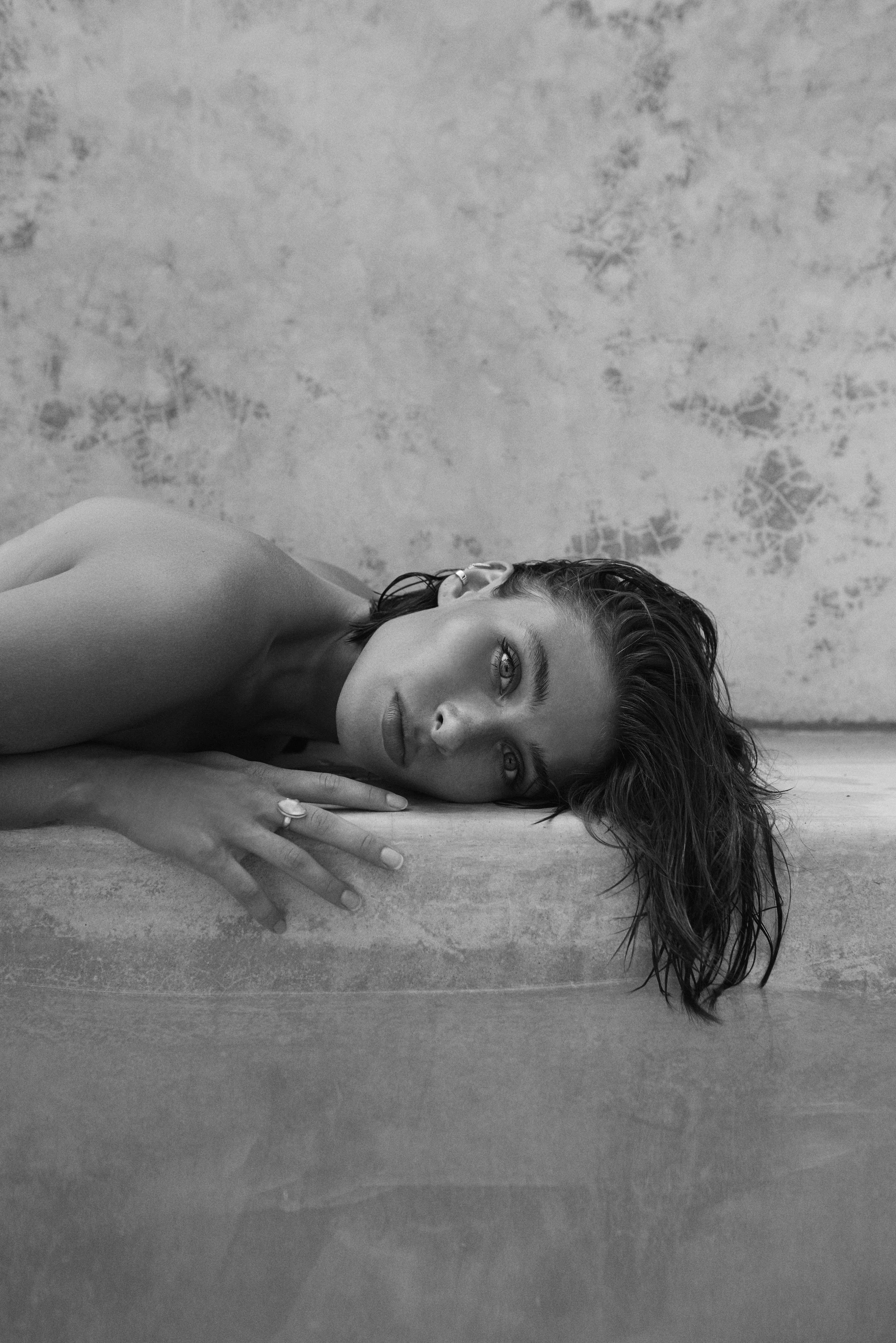 A black and white photo of a woman lying on her side with her head resting on a surface, looking directly at the camera with an intense expression. Her hair is wet and styled to the side, and she wears a ring on her finger and earrings.