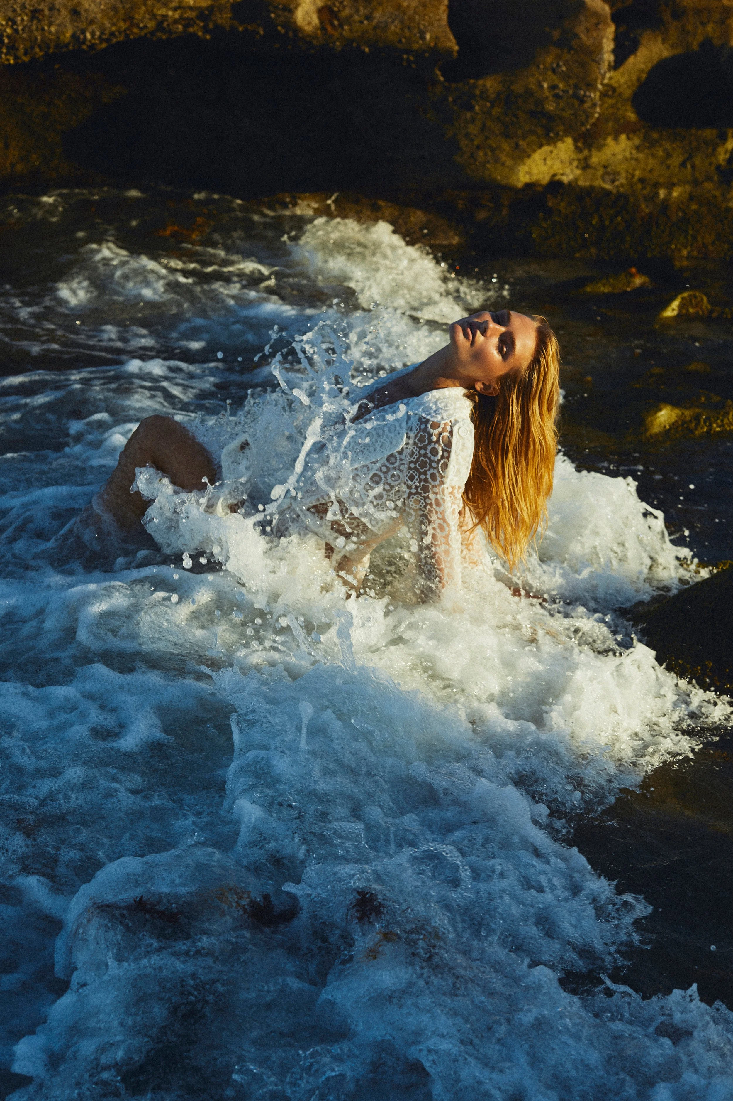 A woman with long blonde hair wearing a white, lacy top is in a rocky ocean, leaning back as waves splash around her during sunset.