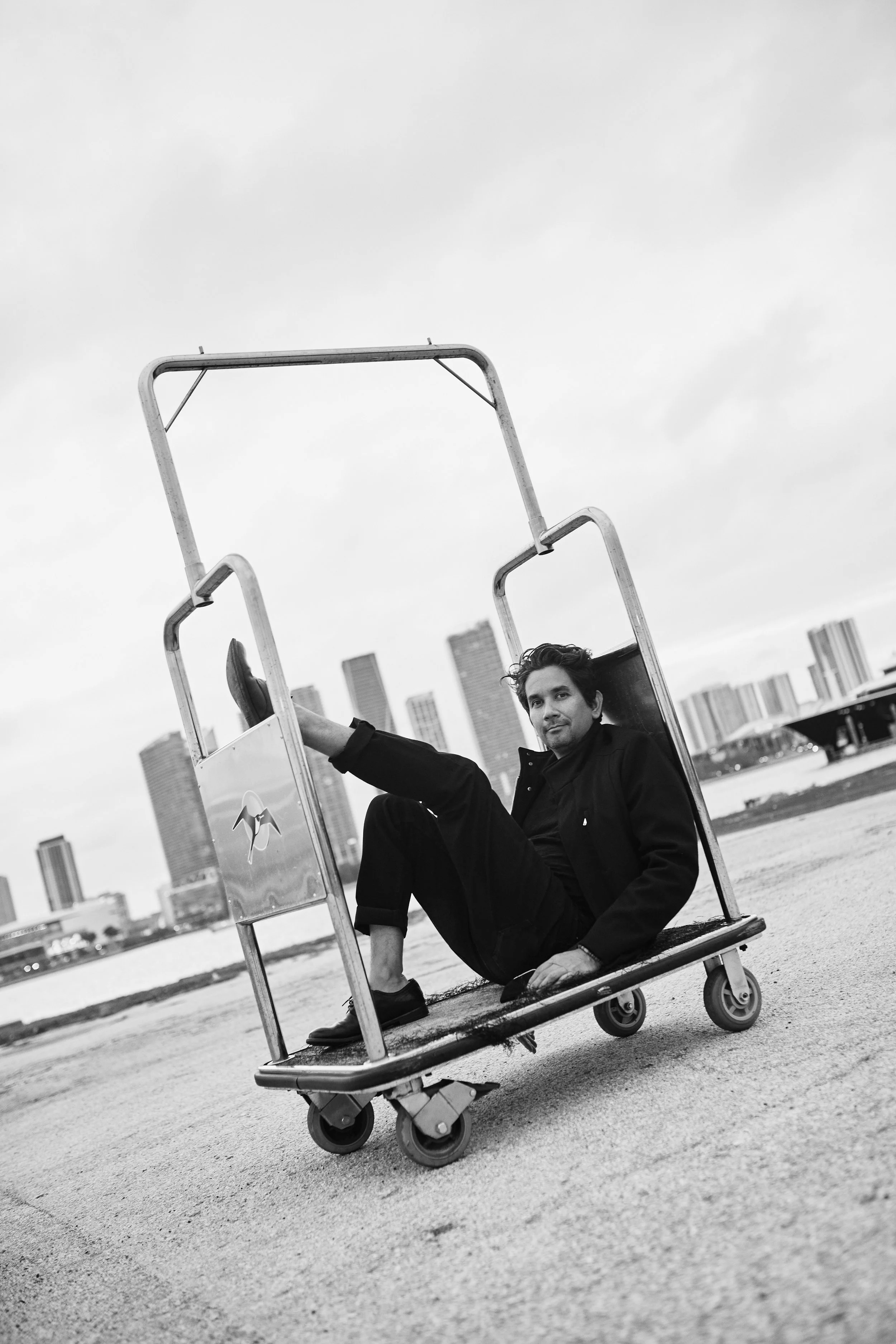 A man sitting on a metal cart with wheels, in an urban setting with tall buildings in the background, photographed in black and white.