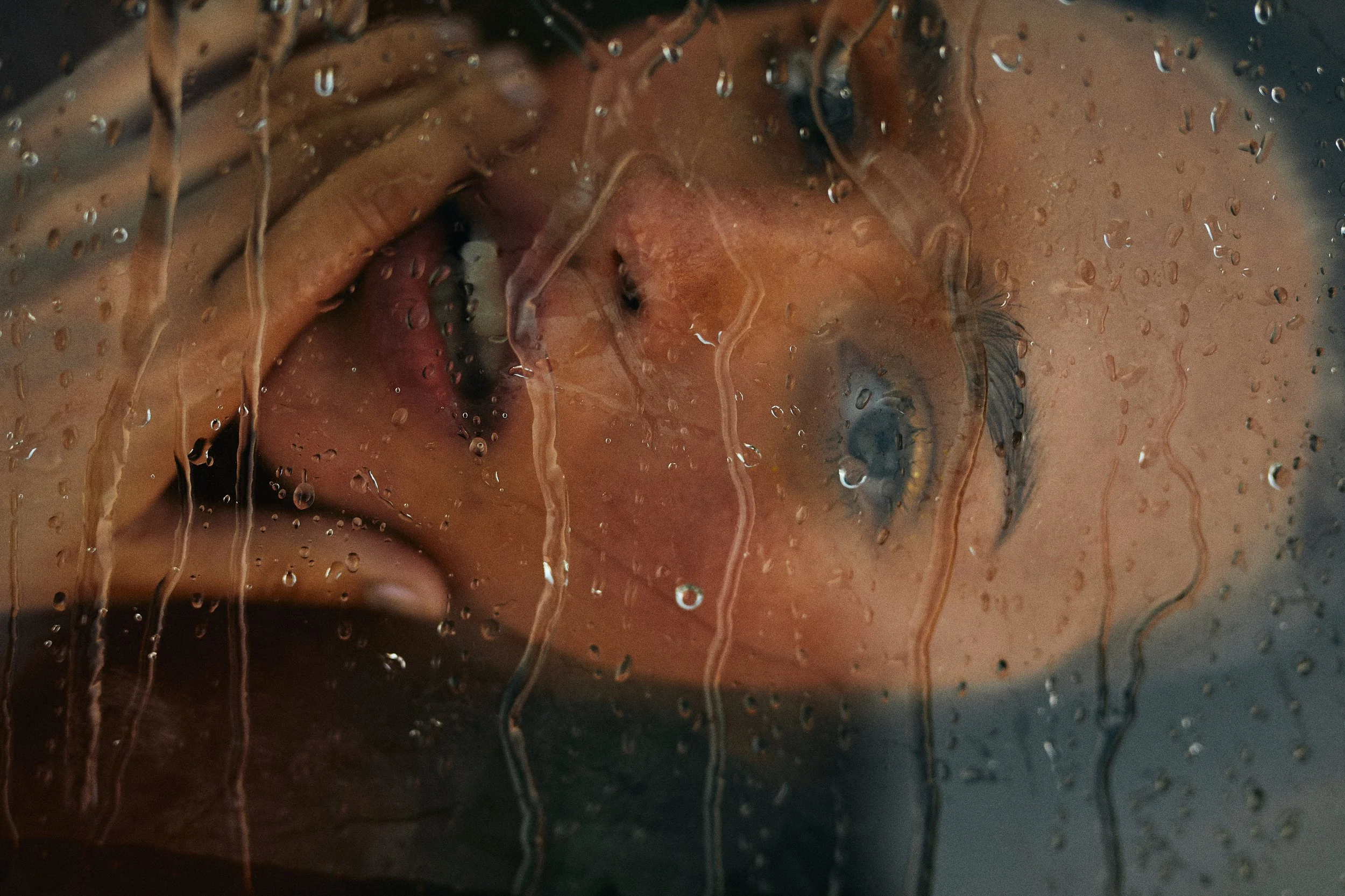 A woman with blue eyes and brown hair looking through a rain-covered glass window, with water streaks obscuring her face.