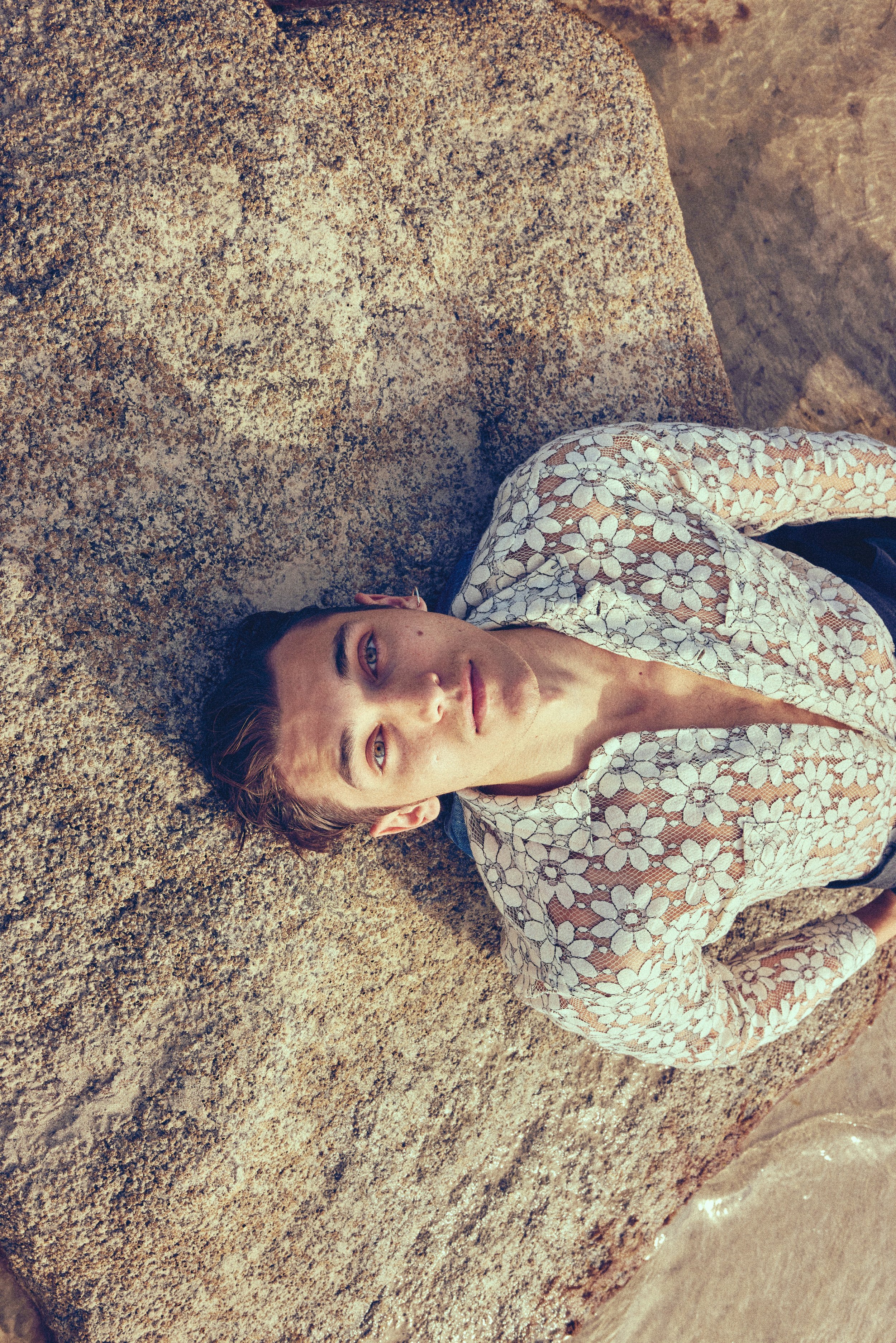 A woman with light skin and blue eyes lying on a sandy surface, wearing a floral lace top, looking at the camera with a calm expression.