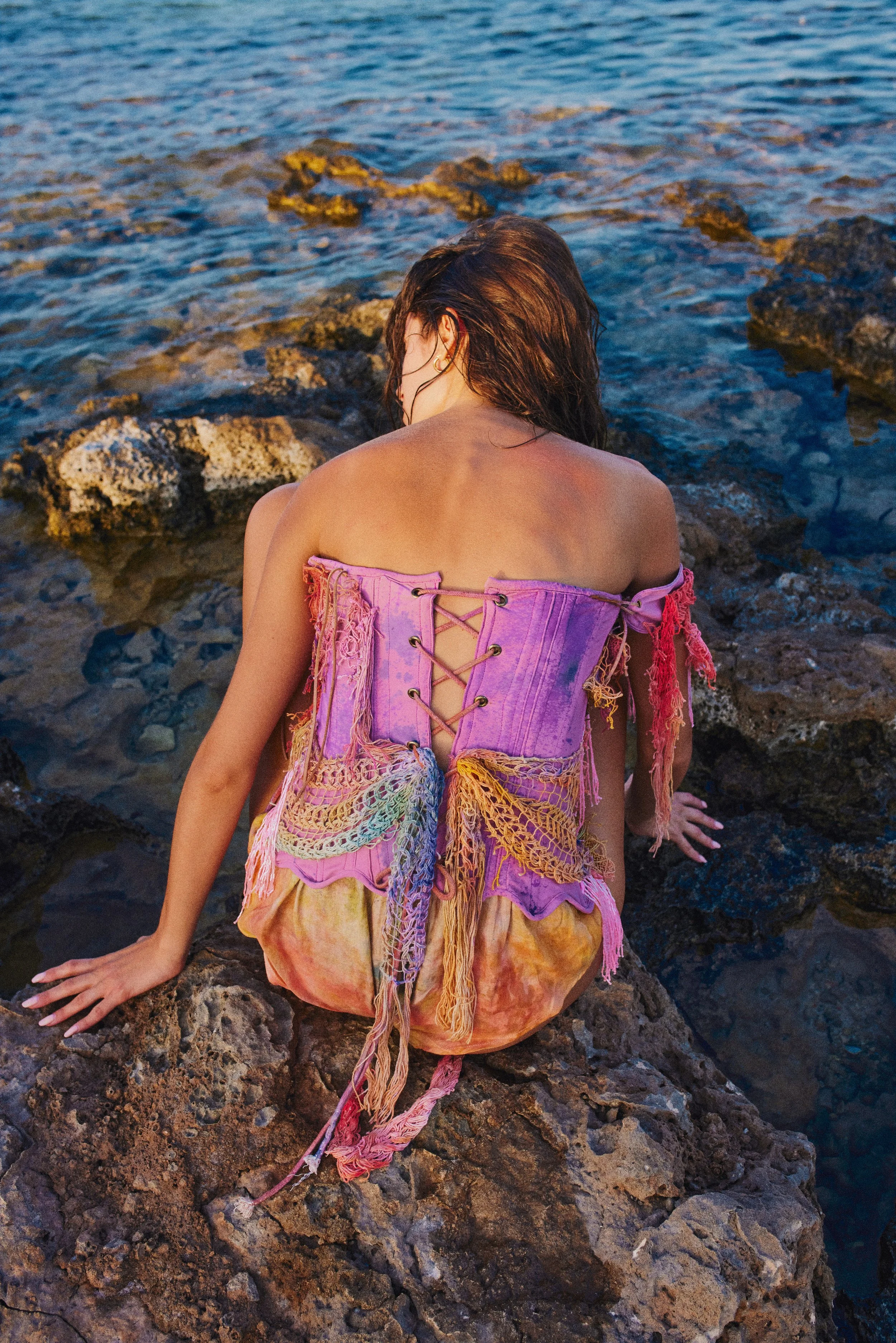 A woman sitting on rocks by the ocean, wearing a colorful, distressed corset top and tie-dye shorts, with the sea and rocks in the background.