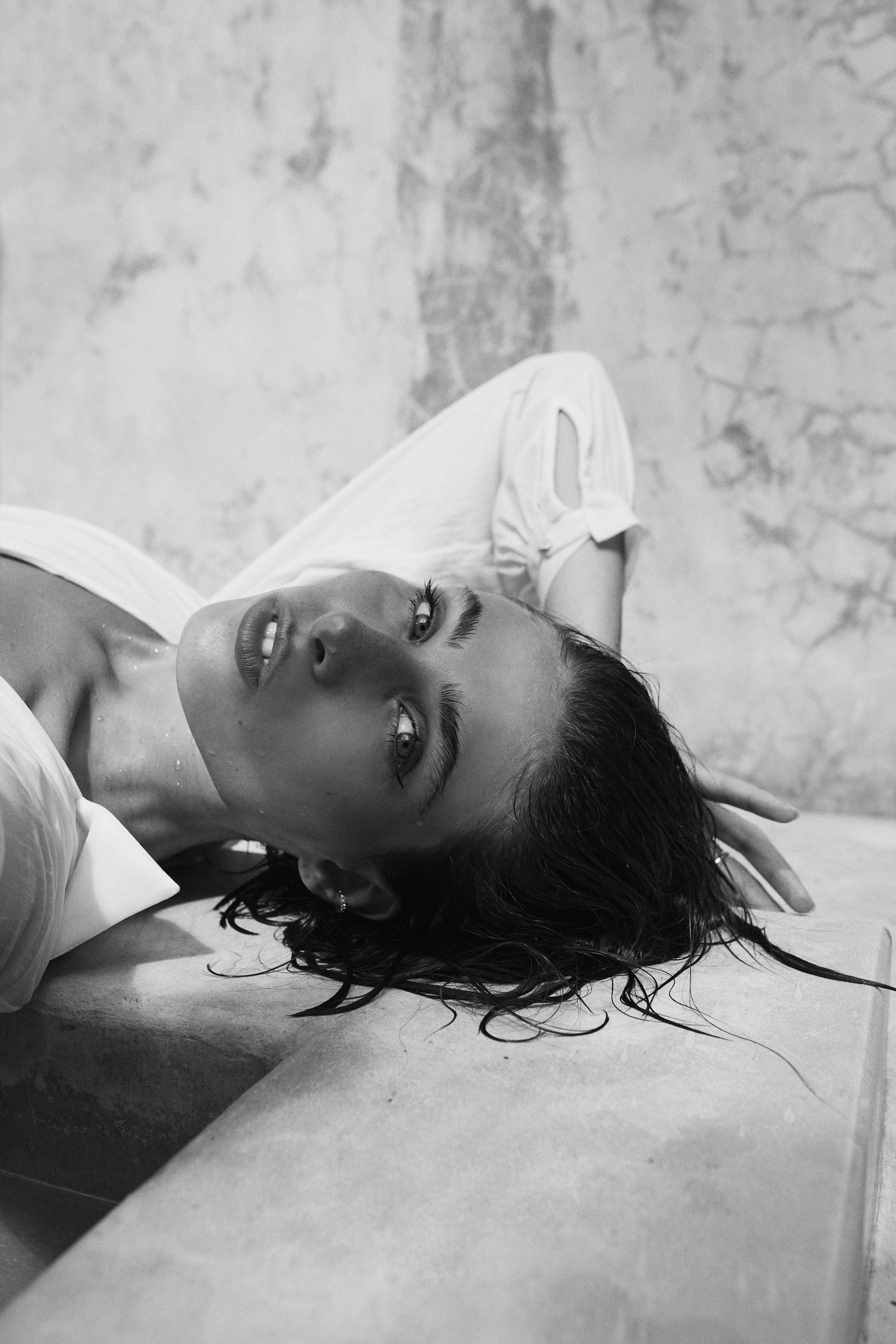 A woman with wet hair lying on a surface, looking up at the camera with an intense expression, in black and white.