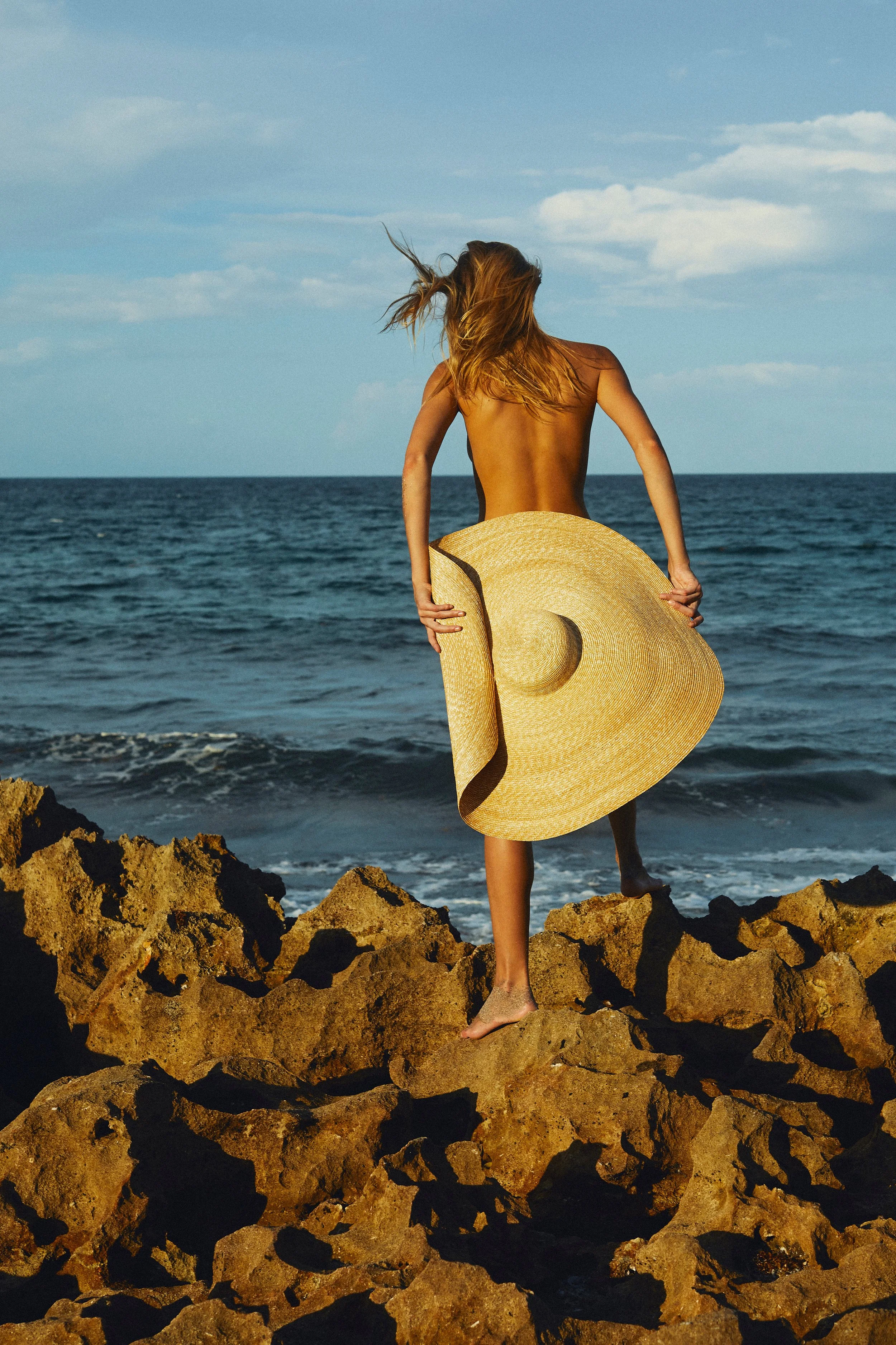 A woman with long hair stands on rocks at the shoreline, holding a large straw sun hat, facing the ocean with her back to the camera on a clear day.