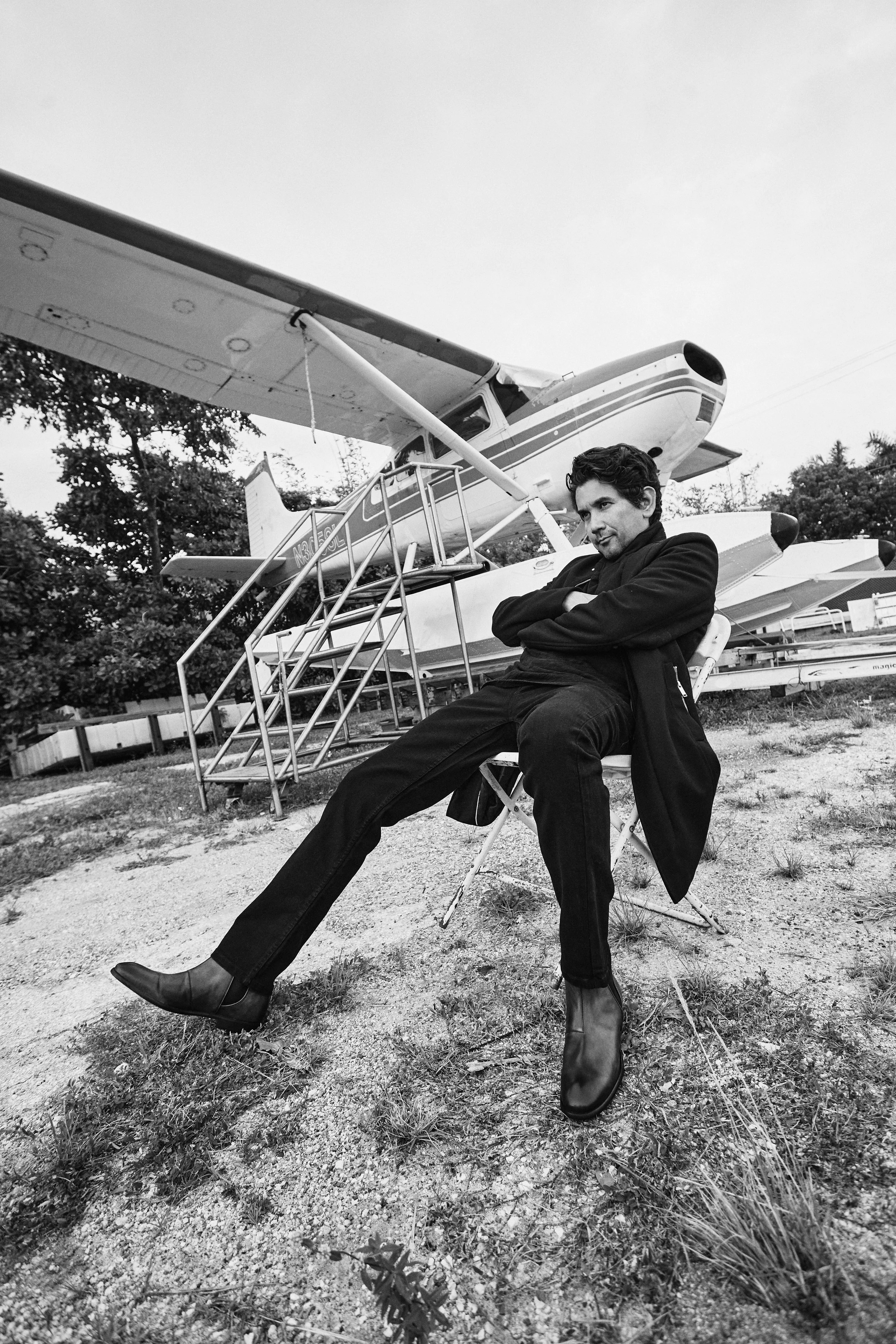 A young man in dark clothing and boots sits with arms crossed and legs stretched out in front of small aircrafts on a grassy area.