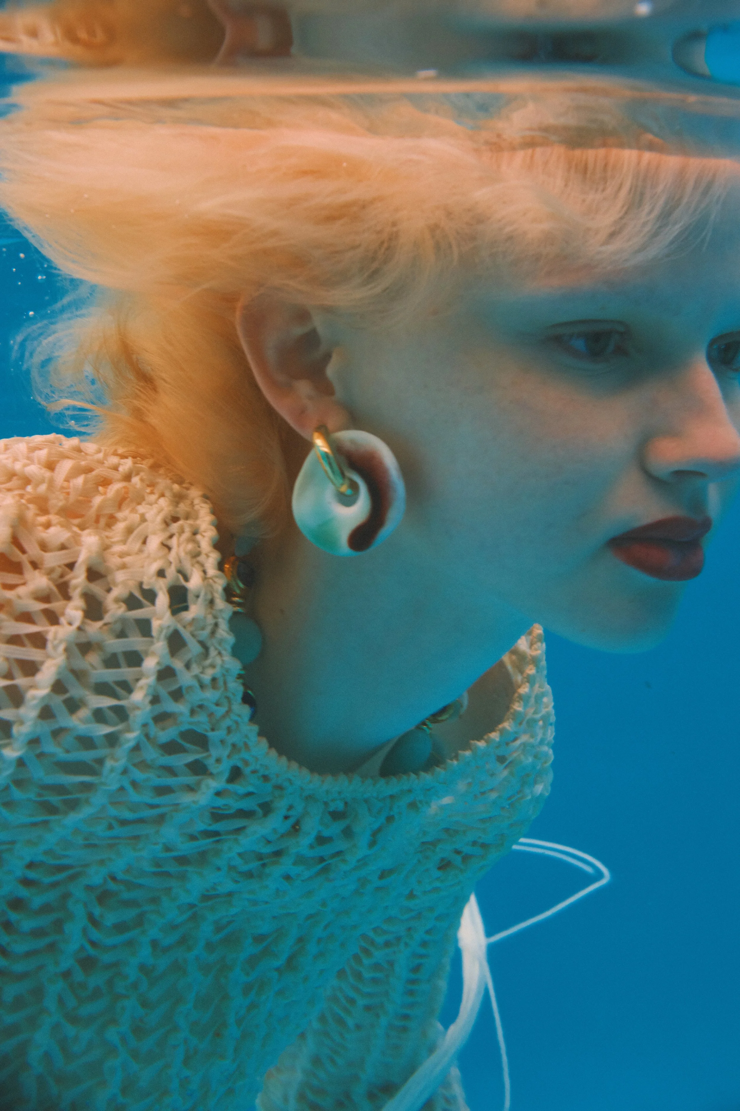 A woman with platinum blonde hair underwater wearing large earrings, a chunky knit sweater, and jewelry.