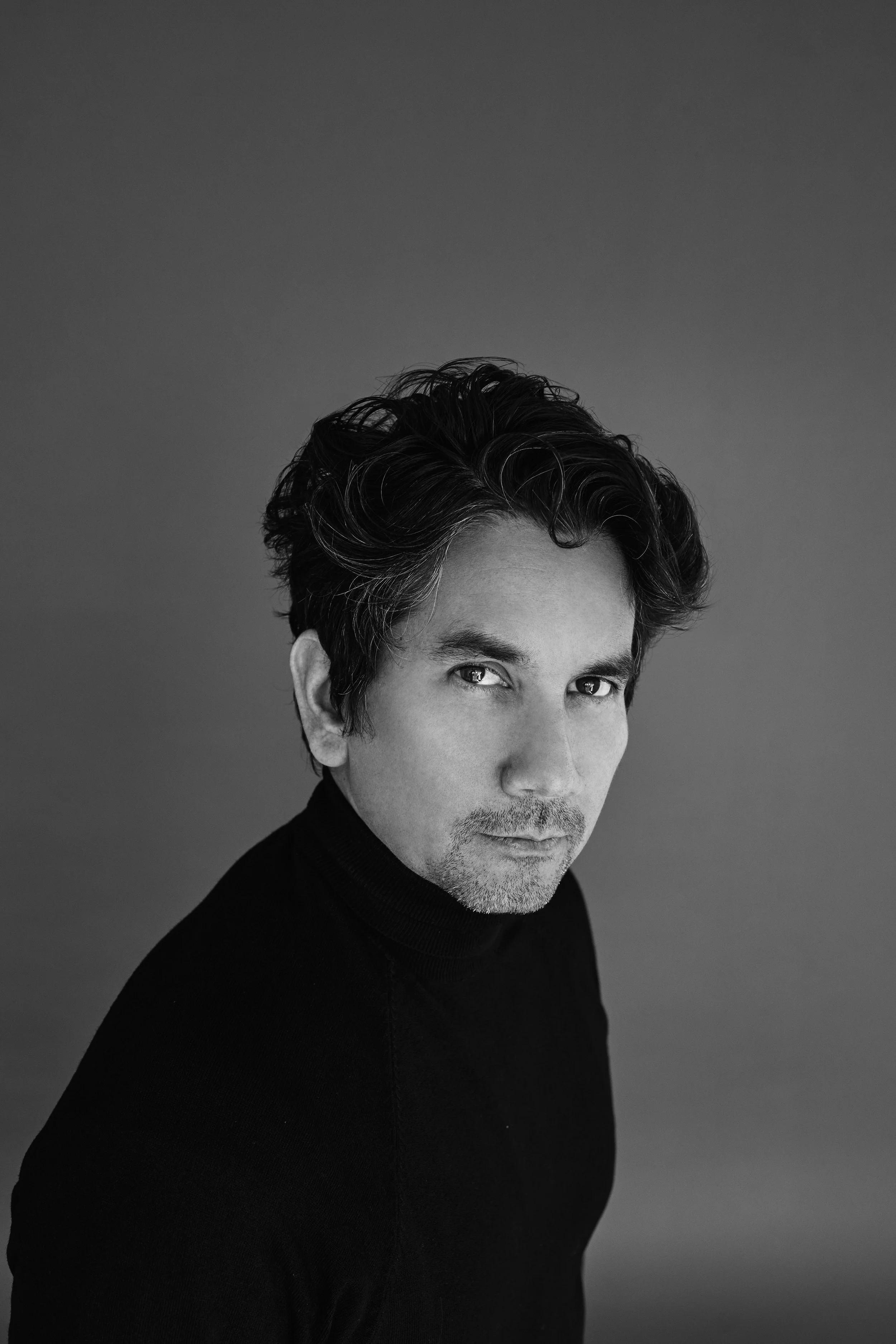 Black and white portrait of a man with dark, wavy hair, wearing a turtleneck sweater, looking directly at the camera with a serious expression.