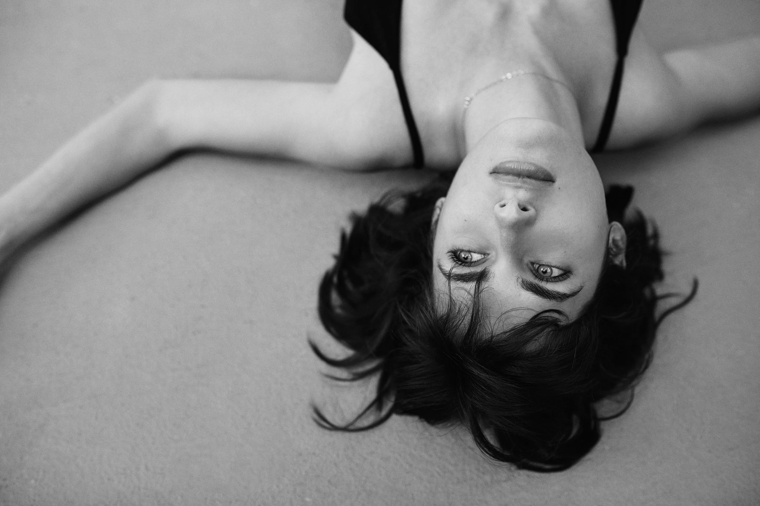 A woman with dark hair lies on her back on a light surface, wearing a black top with thin straps, looking upward with a contemplative expression.