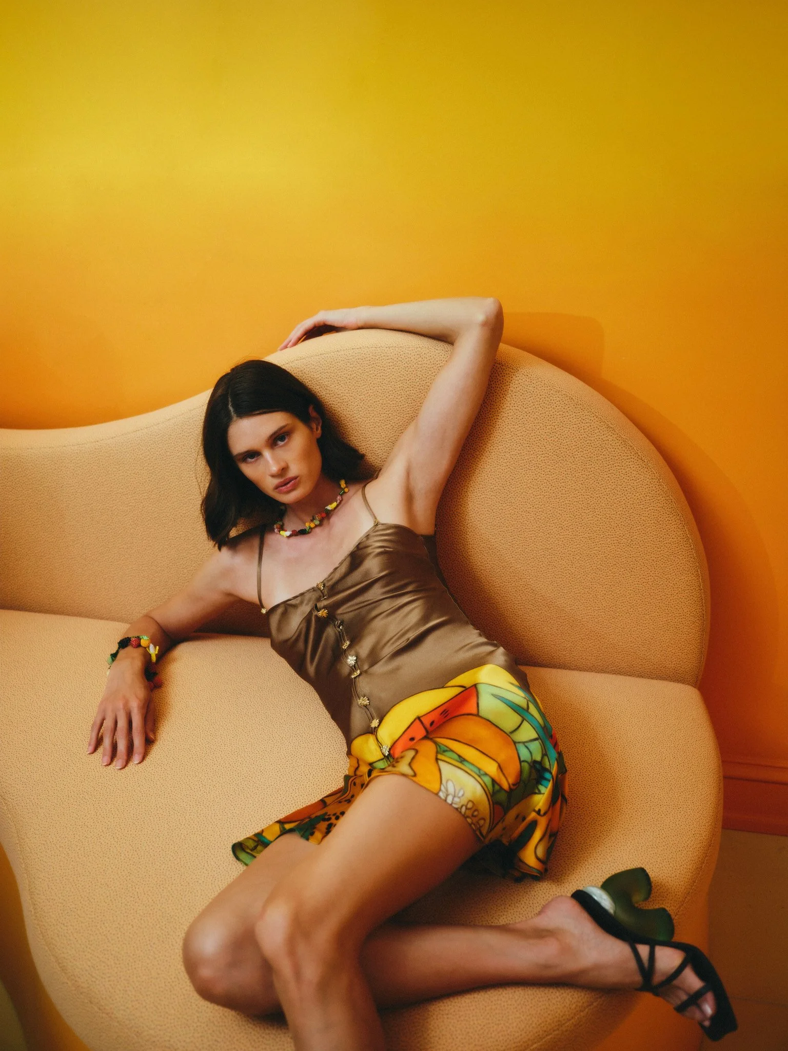 Fashion editorial portrait: woman reclining on a curved beige sofa against a yellow wall, wearing a satin top, colorful fruit-print skirt and beaded jewelry
