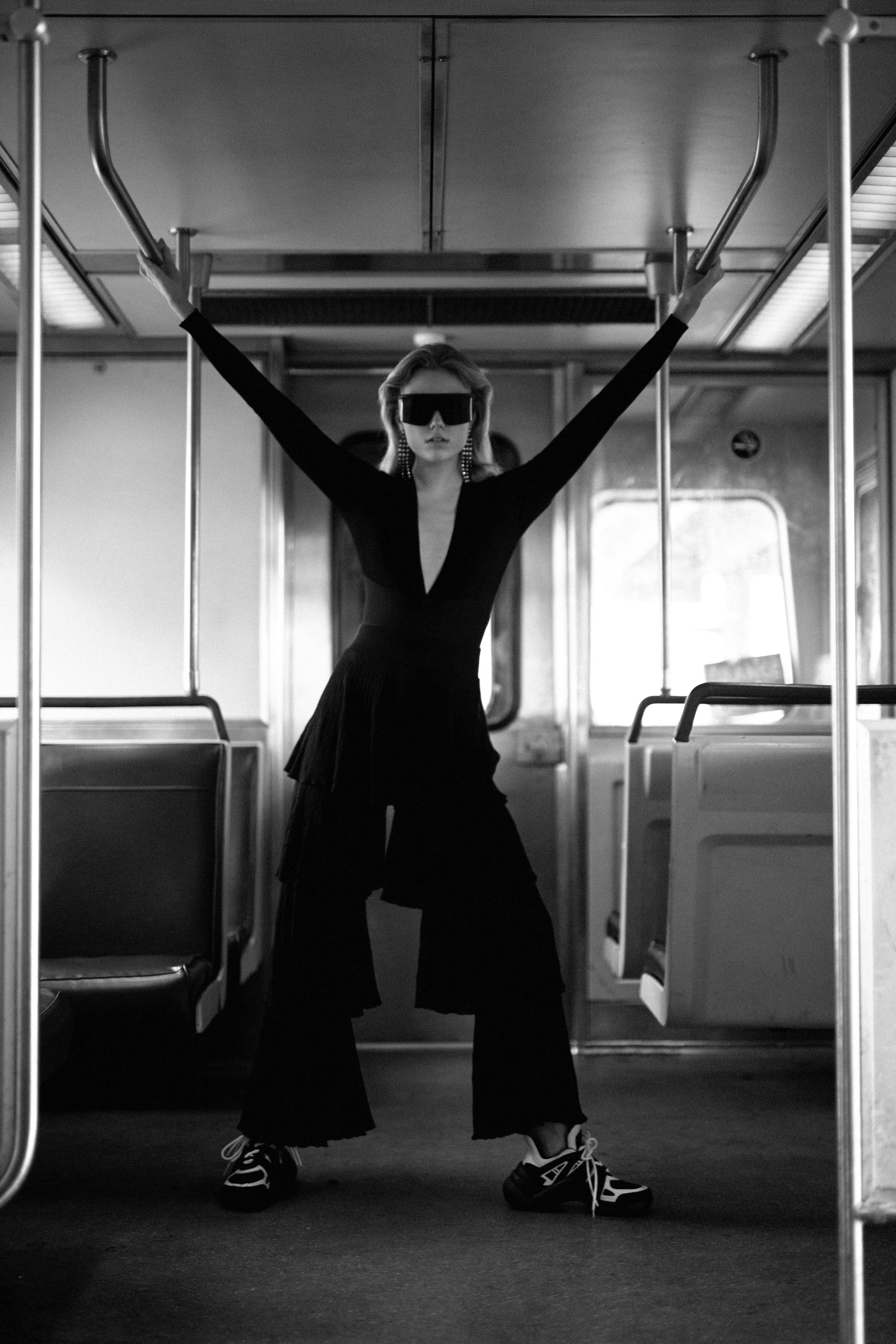 Black and white photo of a woman standing inside a bus, holding onto the overhead handrails with arms extended. She is wearing oversized sunglasses, earrings, a fitted black top, wide-legged black pants, and sneakers.
