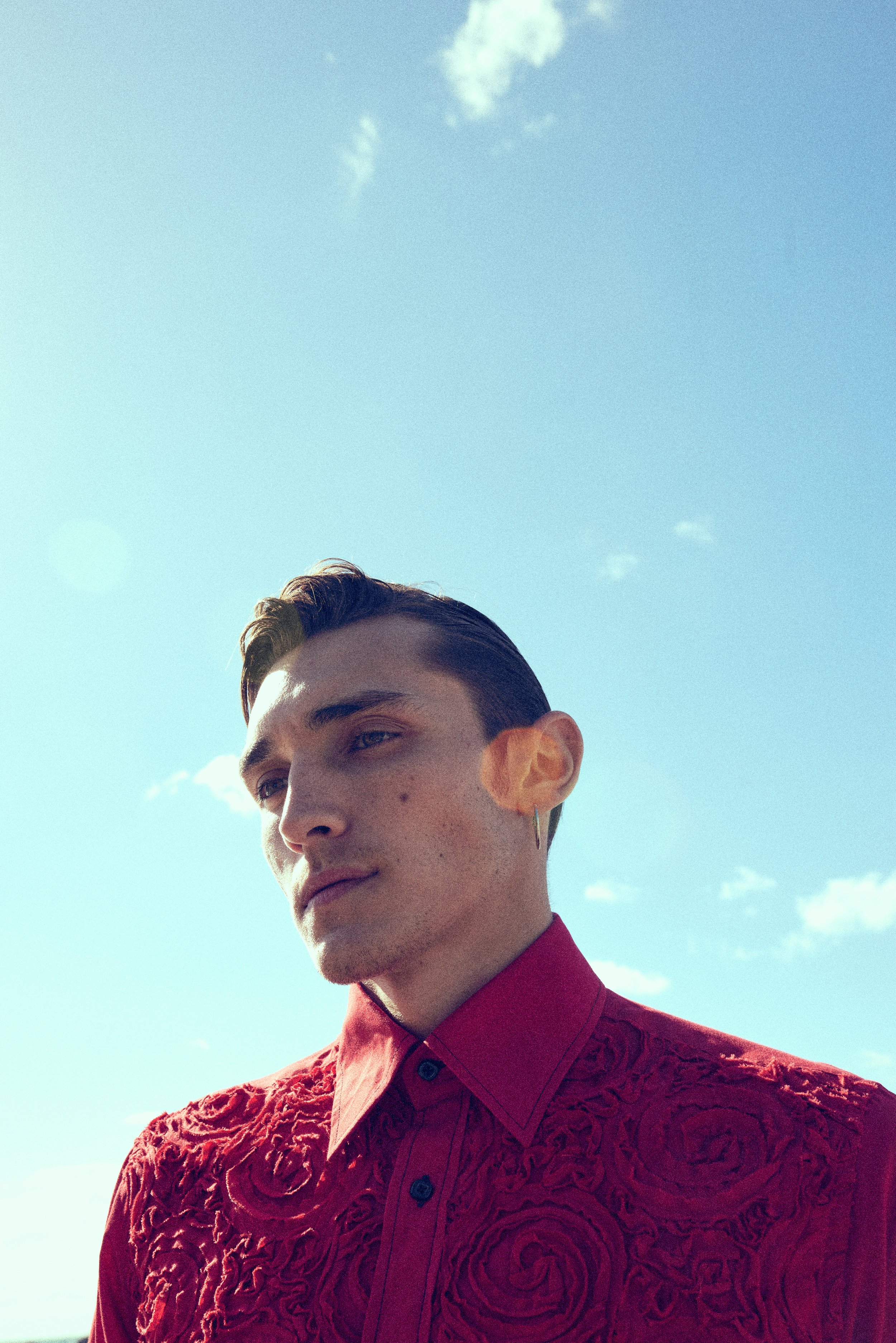 Portrait of a young man with slicked-back hair wearing a red embroidered shirt, standing outdoors against a bright blue sky with scattered white clouds.