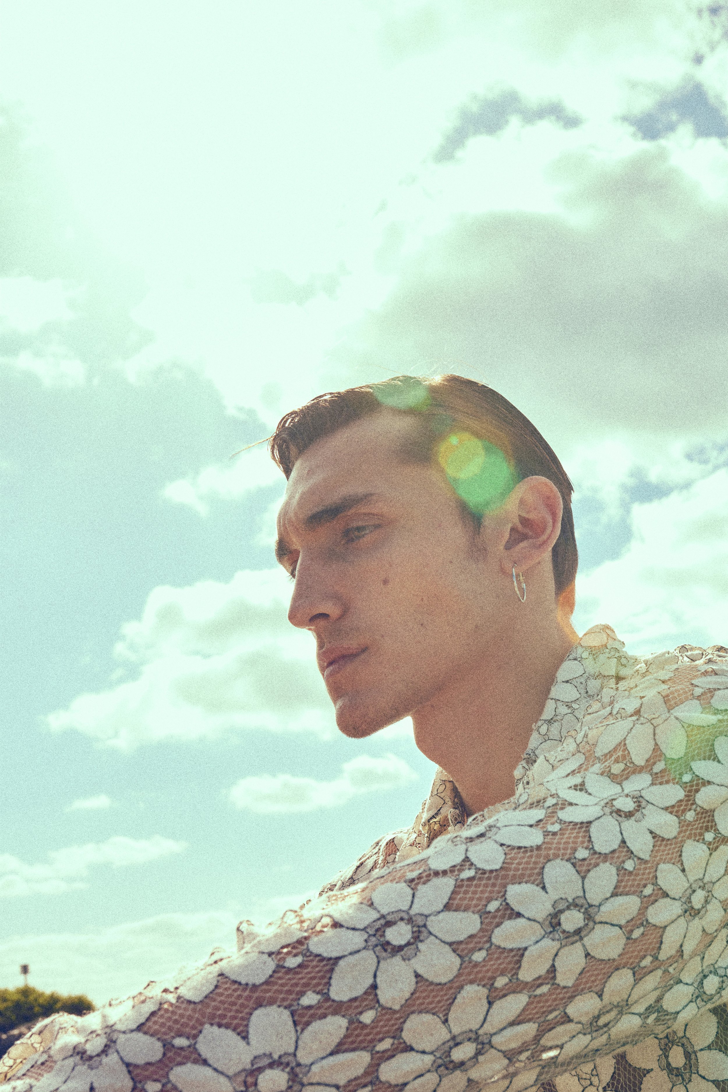 Portrait of a young man with dark hair, earrings, wearing a floral-patterned shirt, outdoors with cloudy sky and sun glare.