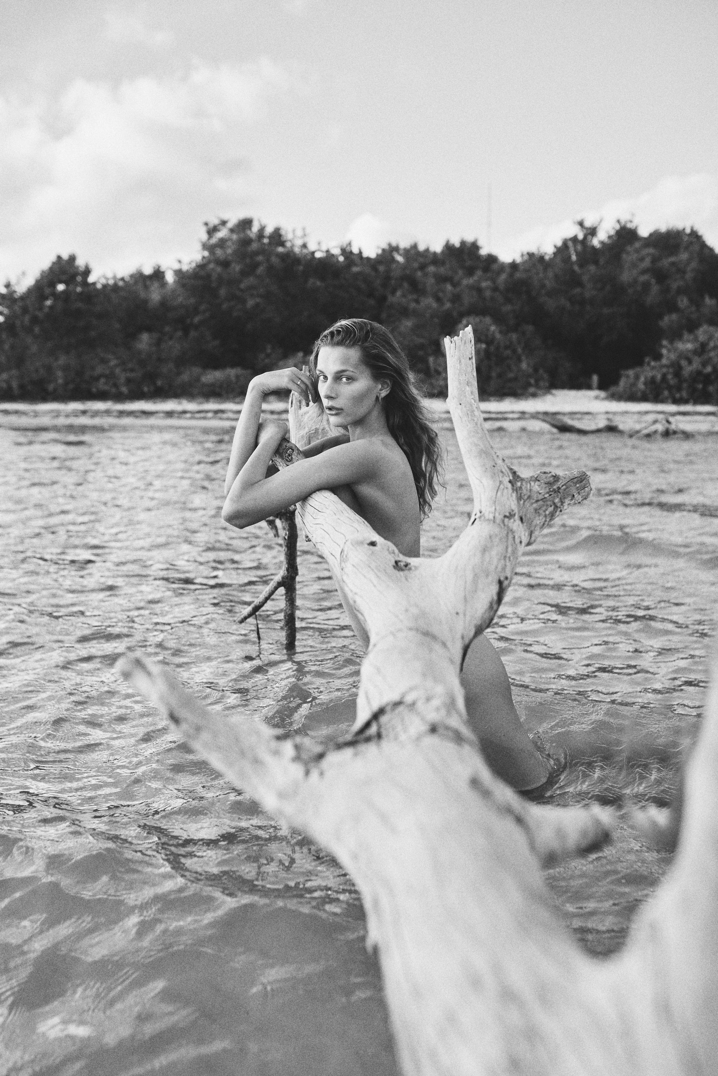 A black-and-white photo of a woman sitting on a piece of driftwood in the water, with trees in the background and clouds in the sky.