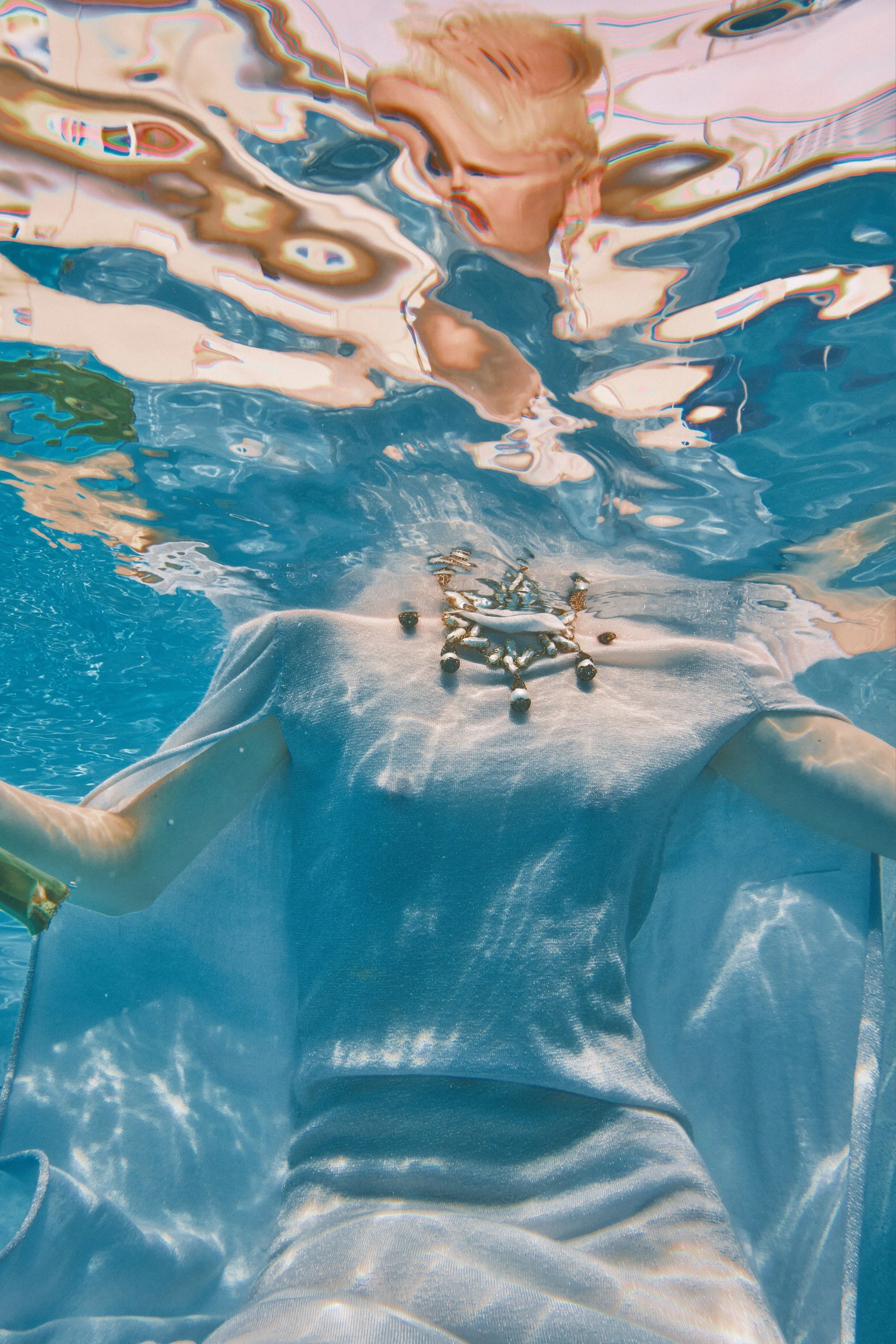 A person underwater wearing a white shirt with a prominent necklace, with the water distorting their face and surroundings.