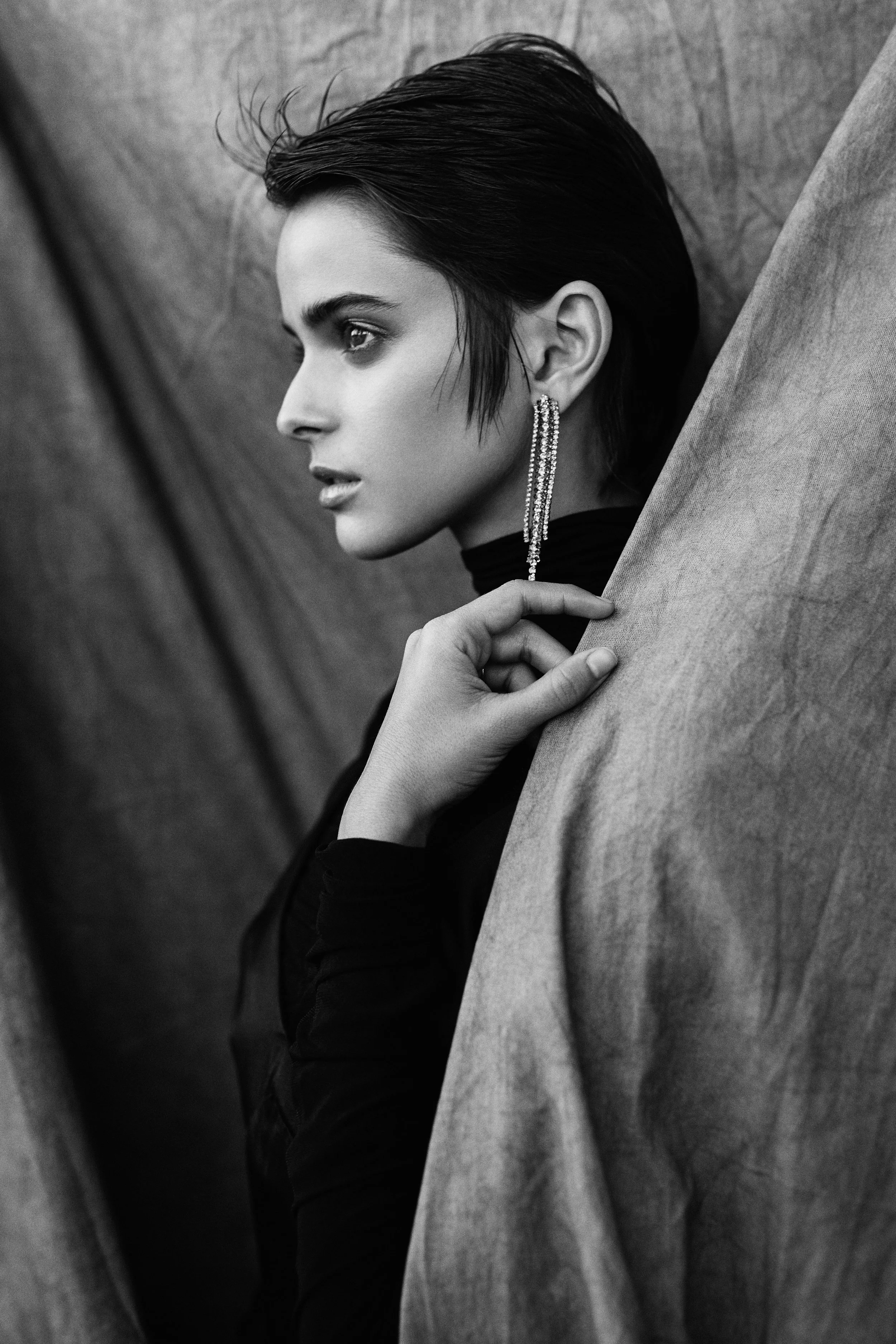 Black and white portrait of a woman with short, sleek hair, wearing long, sparkly earrings and a dark top, gazing sideways with her hand touching her face, against a textured fabric backdrop.