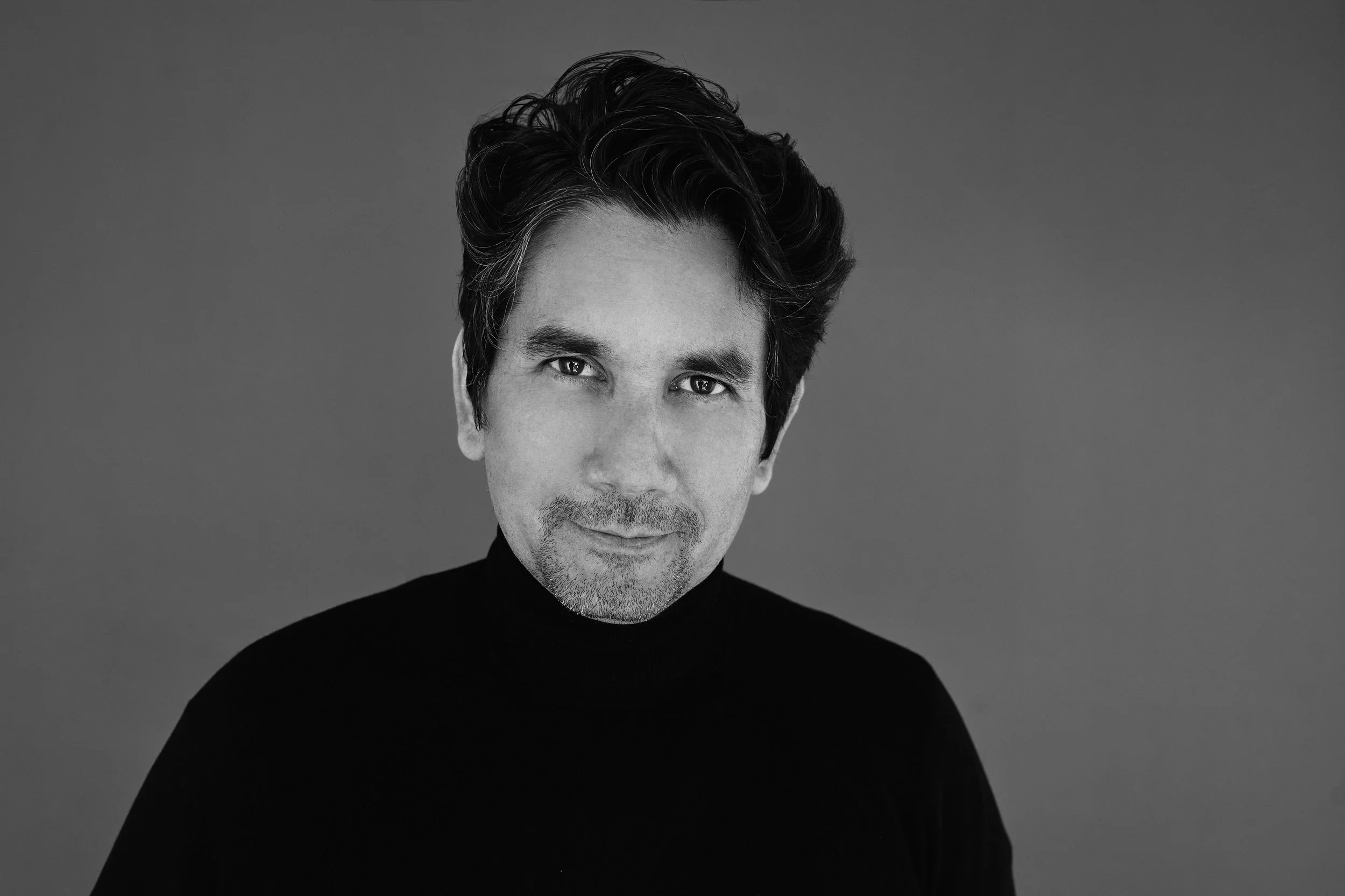 Black and white portrait of a man with dark, wavy hair, wearing a black turtleneck, smiling slightly and looking at the camera.