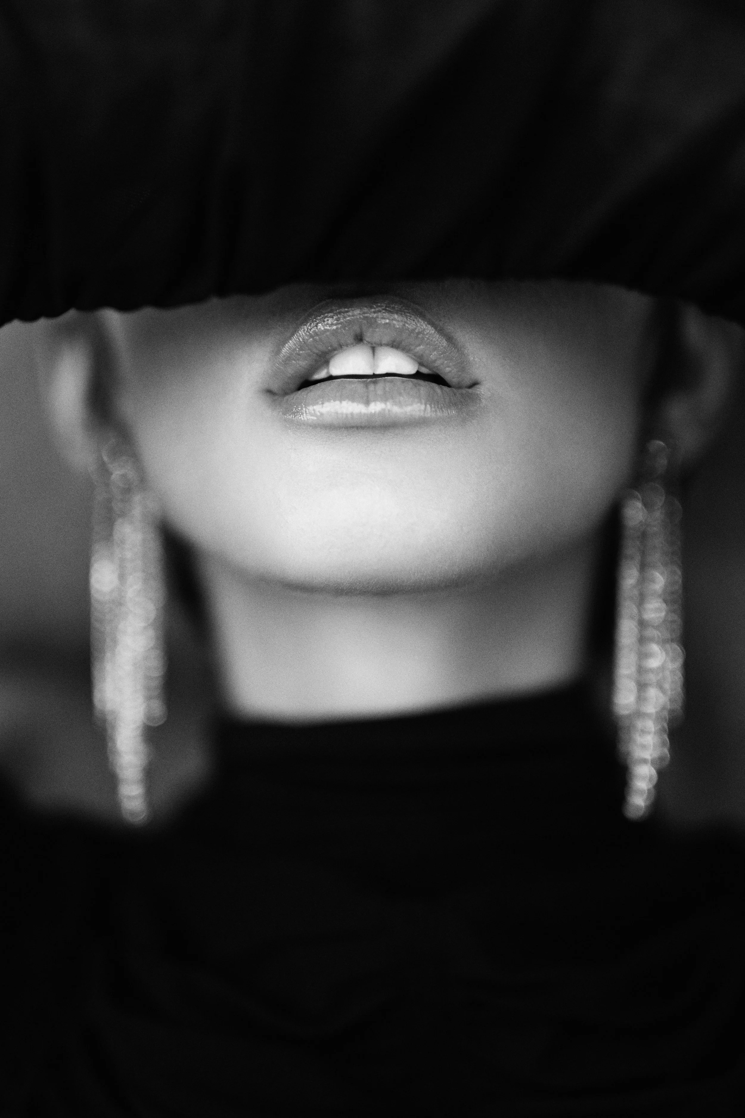 Close-up black and white photograph of a woman's face partially obscured by a dark hat, showing lips and dangling earrings.