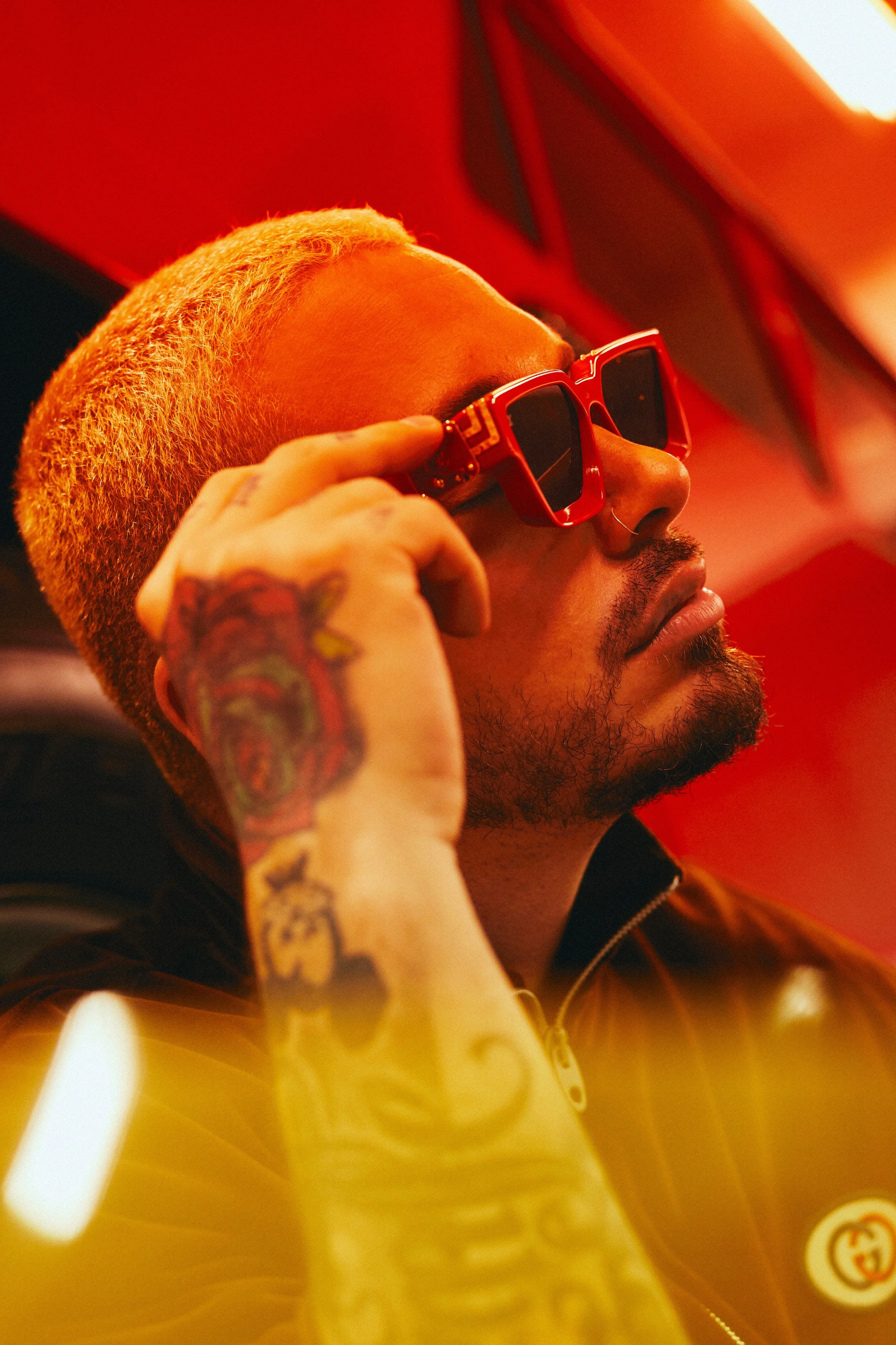 A man with a shiny blond buzz cut, wearing red patterned sunglasses, a gold Gucci jacket, and a nose ring, is posing against a red background.