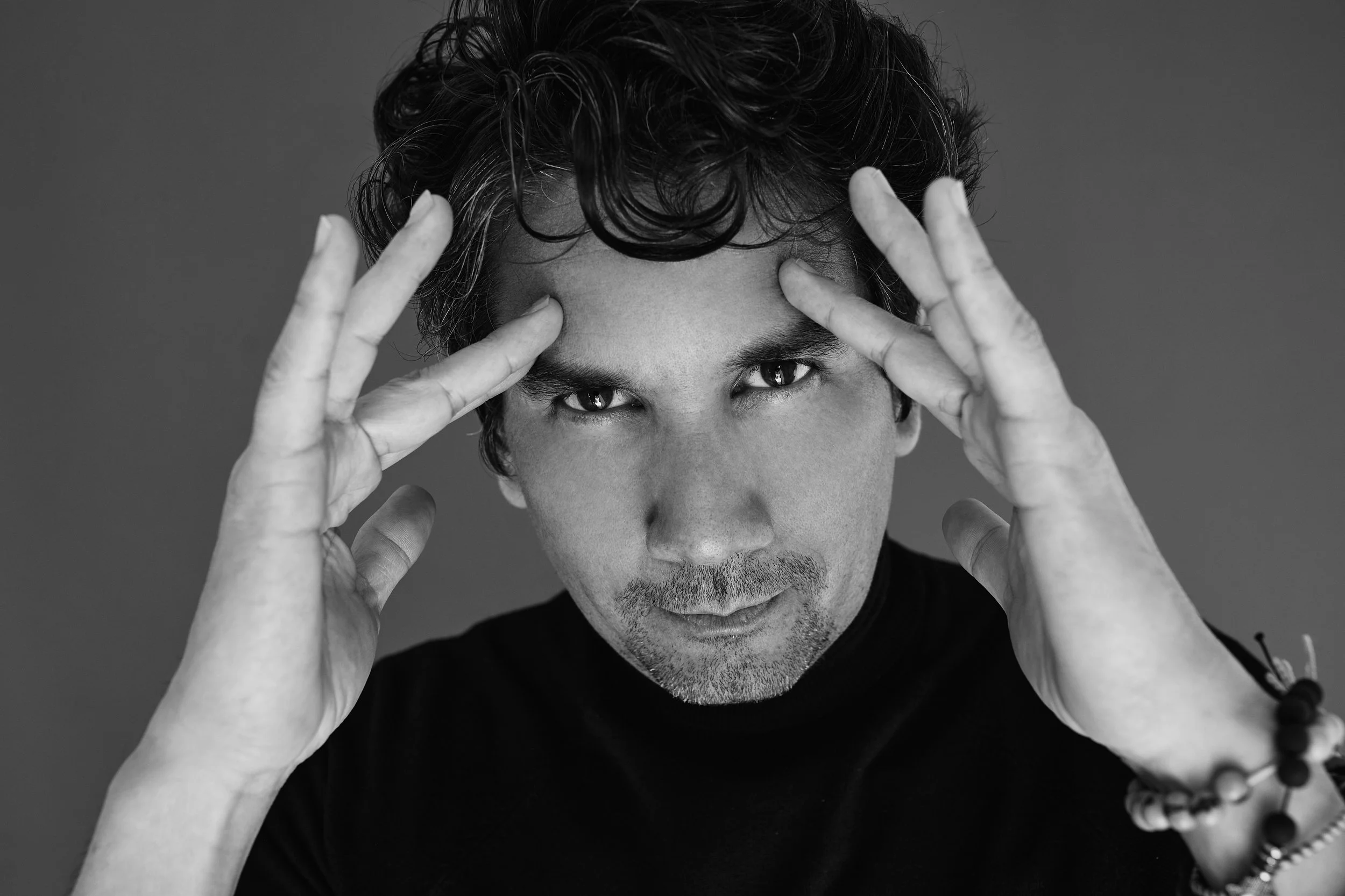 Black and white photo of a man with wavy hair staring intensely at the camera, holding his hands near his head in a thoughtful pose, wearing a black shirt and beaded bracelet.