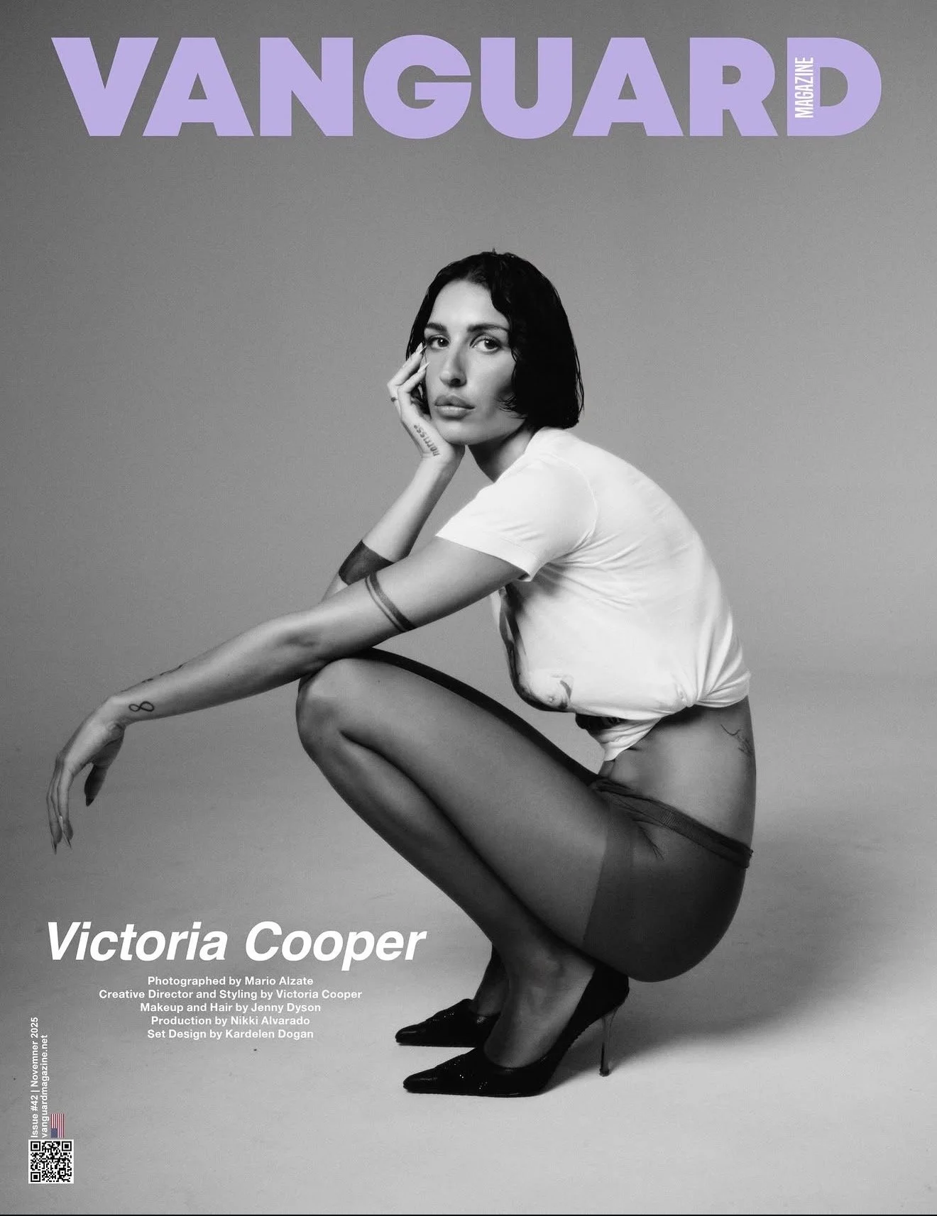 Black and white magazine cover featuring Victoria Cooper in a crouching pose, wearing a t-shirt, shorts, stockings, and high heels, with her hand resting on her face and her other arm extended downward. The title 'Vanguard Magazine' is at the top, an