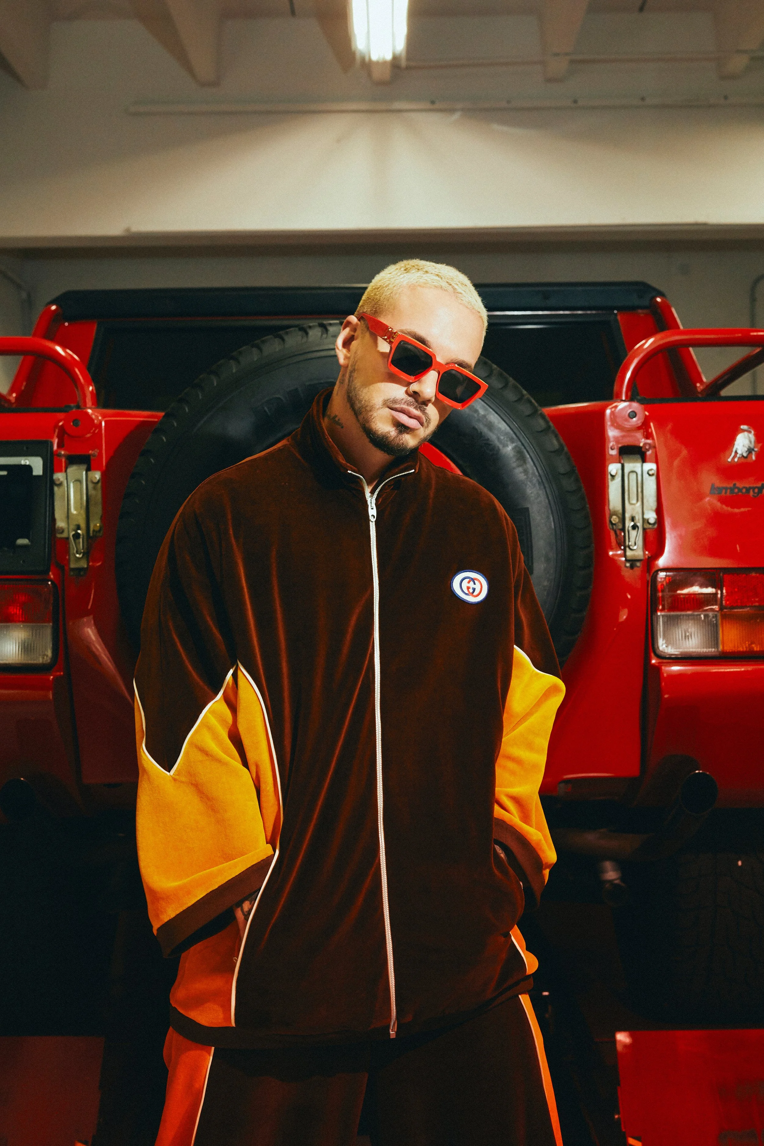 Young man with blonde hair and sunglasses standing in front of a red Lamborghini in a garage.