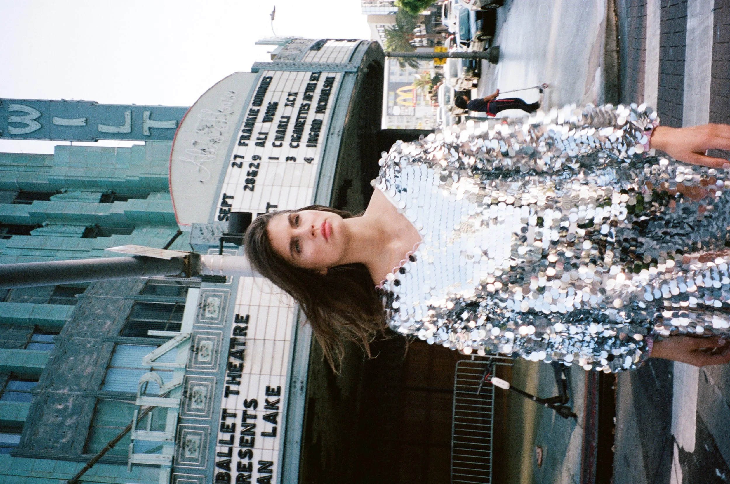 A woman in a shiny, sequined dress standing in front of a theater marquee on a city street.