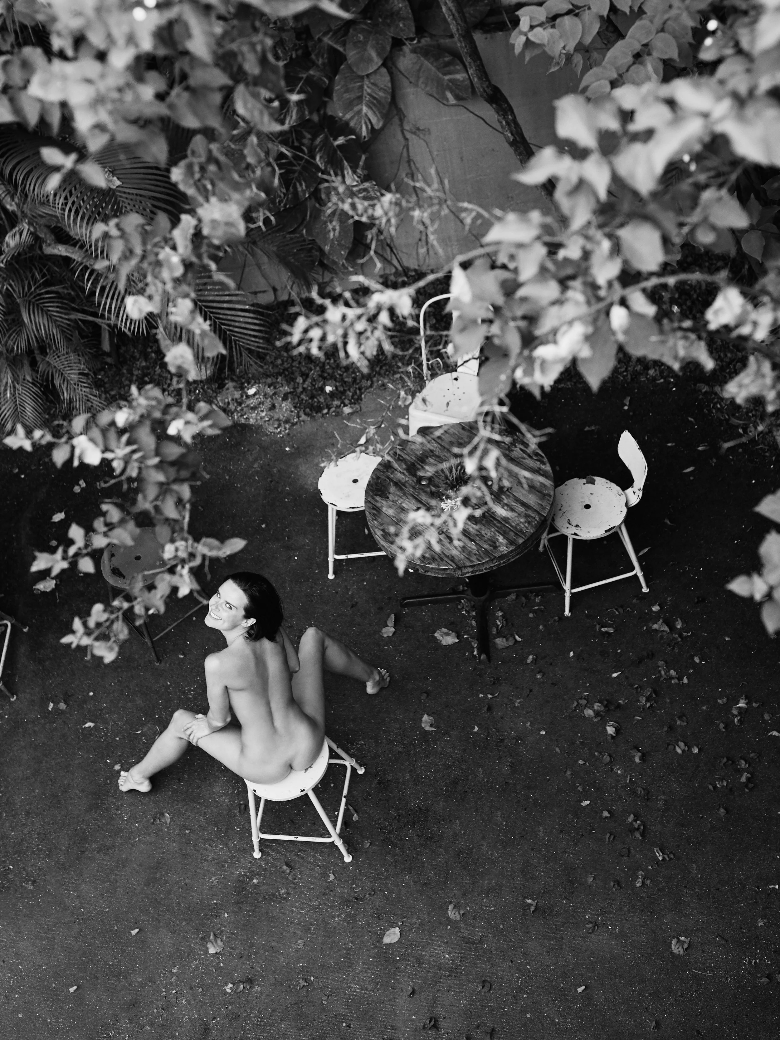 Black and white photo of a topless woman sitting on a chair outdoors, looking up and smiling, with trees and a table and chairs in the background.