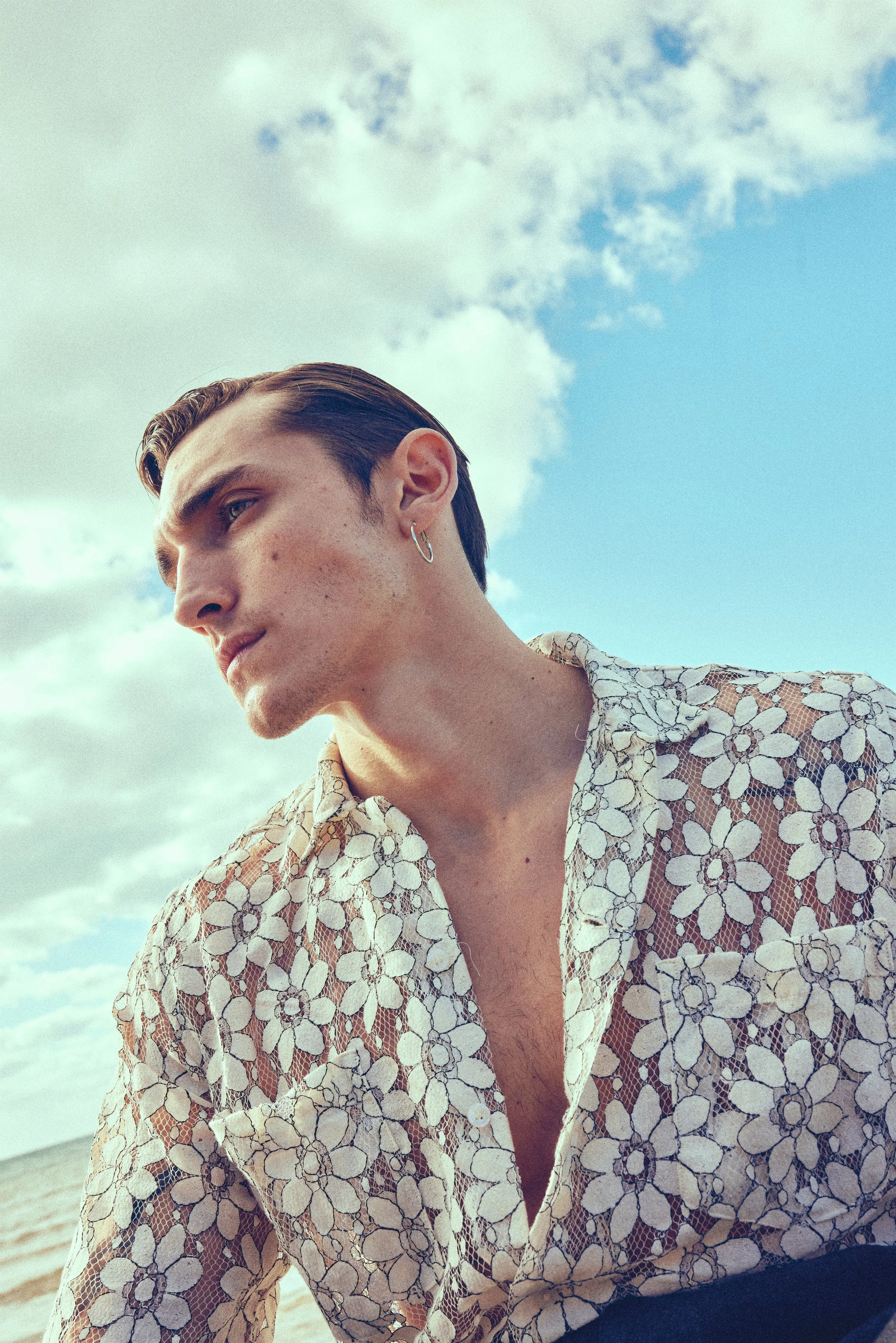 A young man with dark slicked-back hair, wearing a floral lace shirt, looking to the side against a cloudy sky background.
