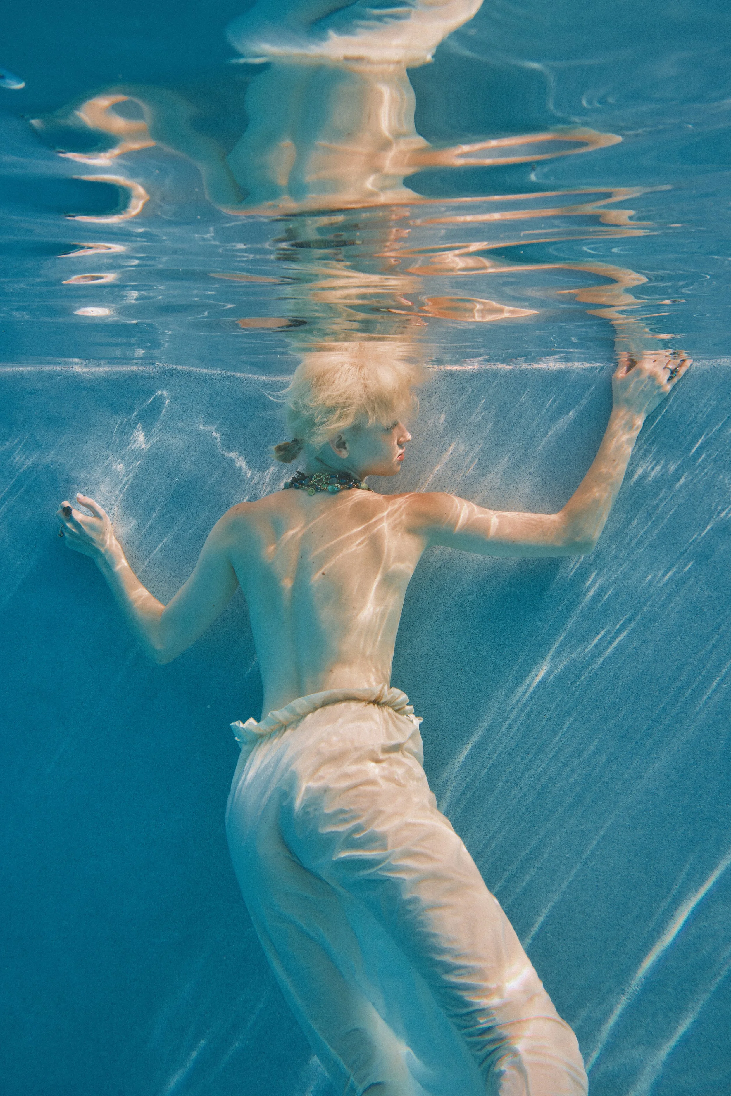 A person with blonde hair swimming underwater in a pool, wearing light-colored pants and a necklace, with the water's surface and reflections visible above.