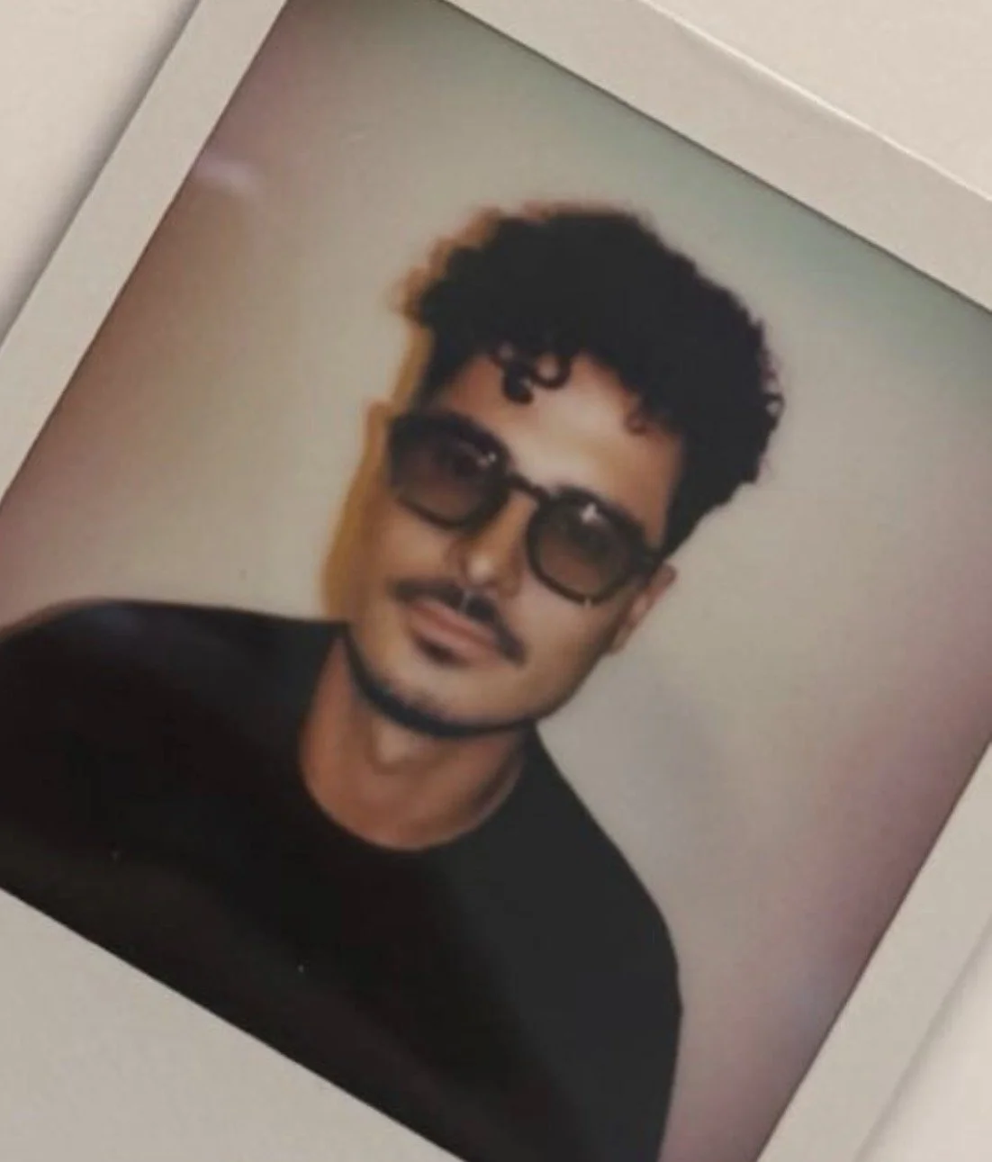 Photograph of a young man with curly black hair, sunglasses, and a goatee, wearing a black shirt, smiling at the camera.