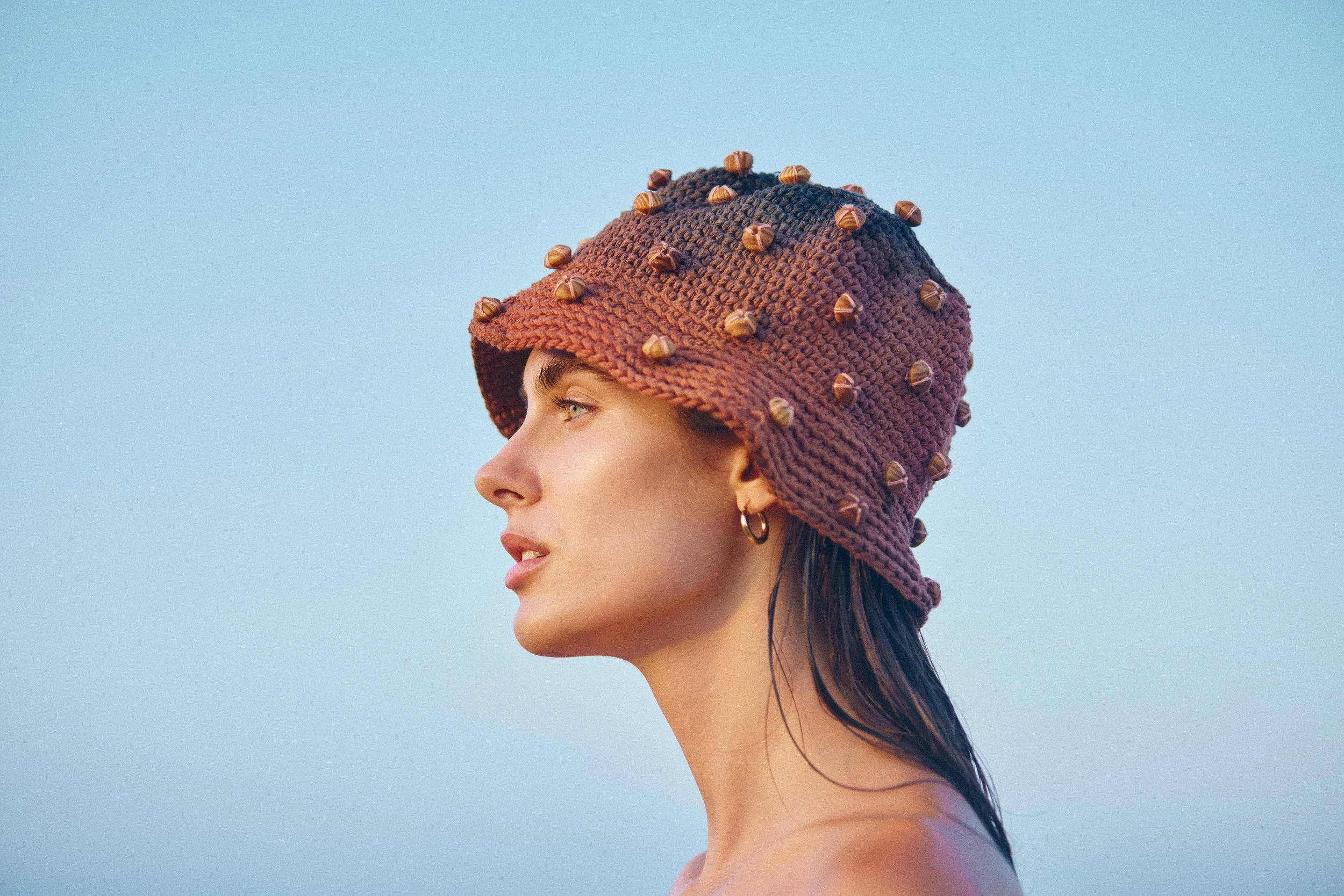 A woman in profile wearing a purple knit hat decorated with small nuts, against a clear blue sky.