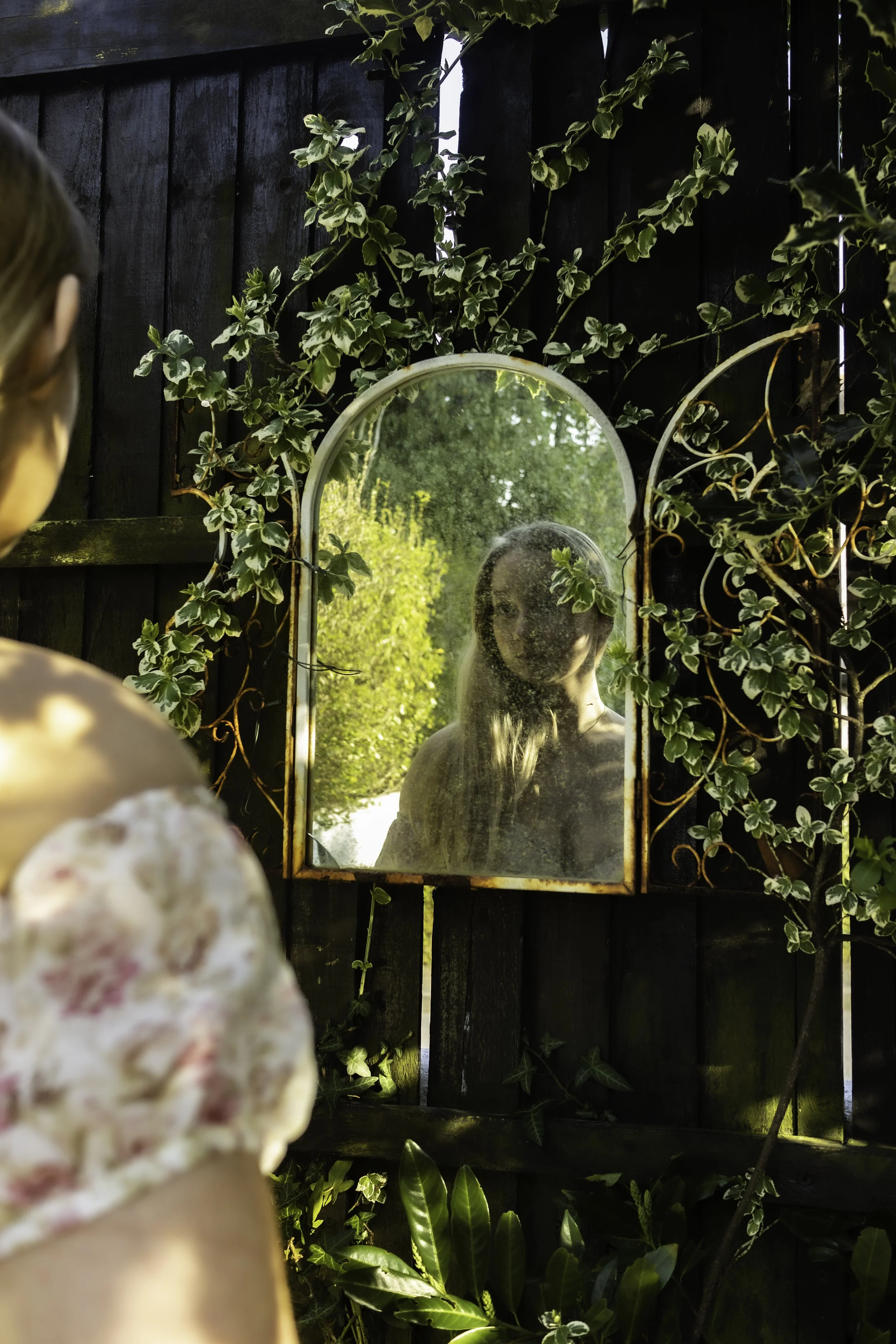 A young girl with floral clothing and a floral headband looks into a small, rounded mirror attached to a black wooden fence. The mirror reflects her face and the surrounding greenery, including various plants and vines.