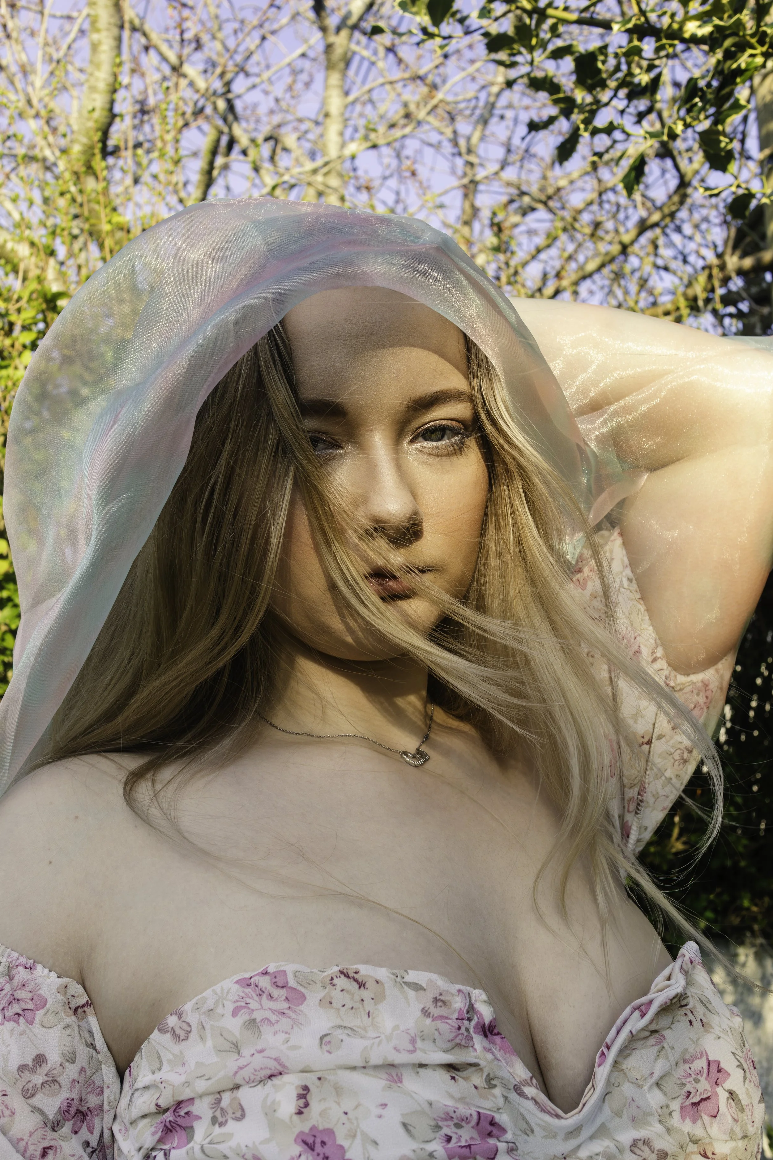 A woman outdoors with sunlight on her face, wearing a floral off-shoulder dress and a sheer, iridescent shawl, with trees and blue sky in the background.