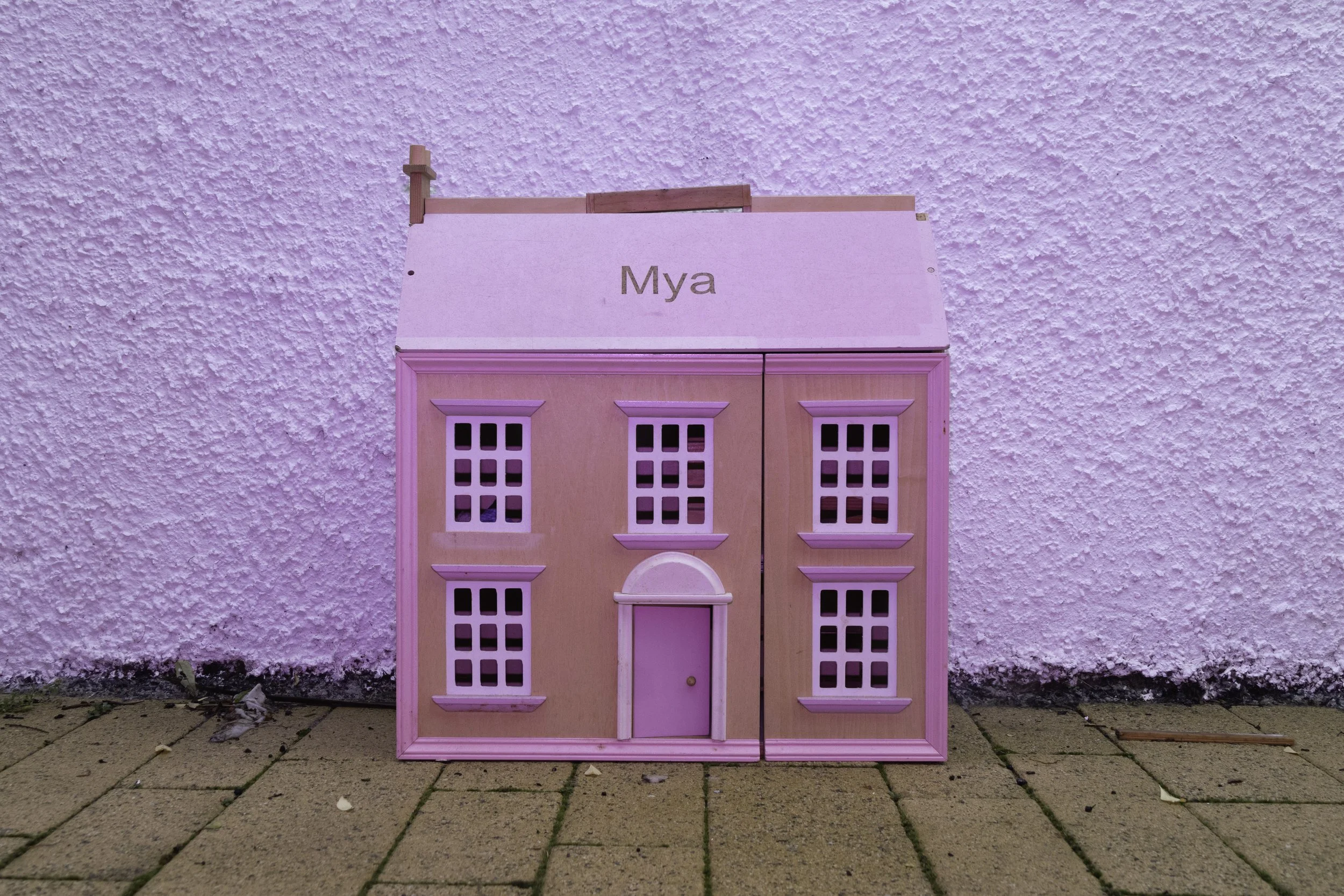 Pink dollhouse with white windows and a door, set against a textured white wall on a paved outdoor surface.