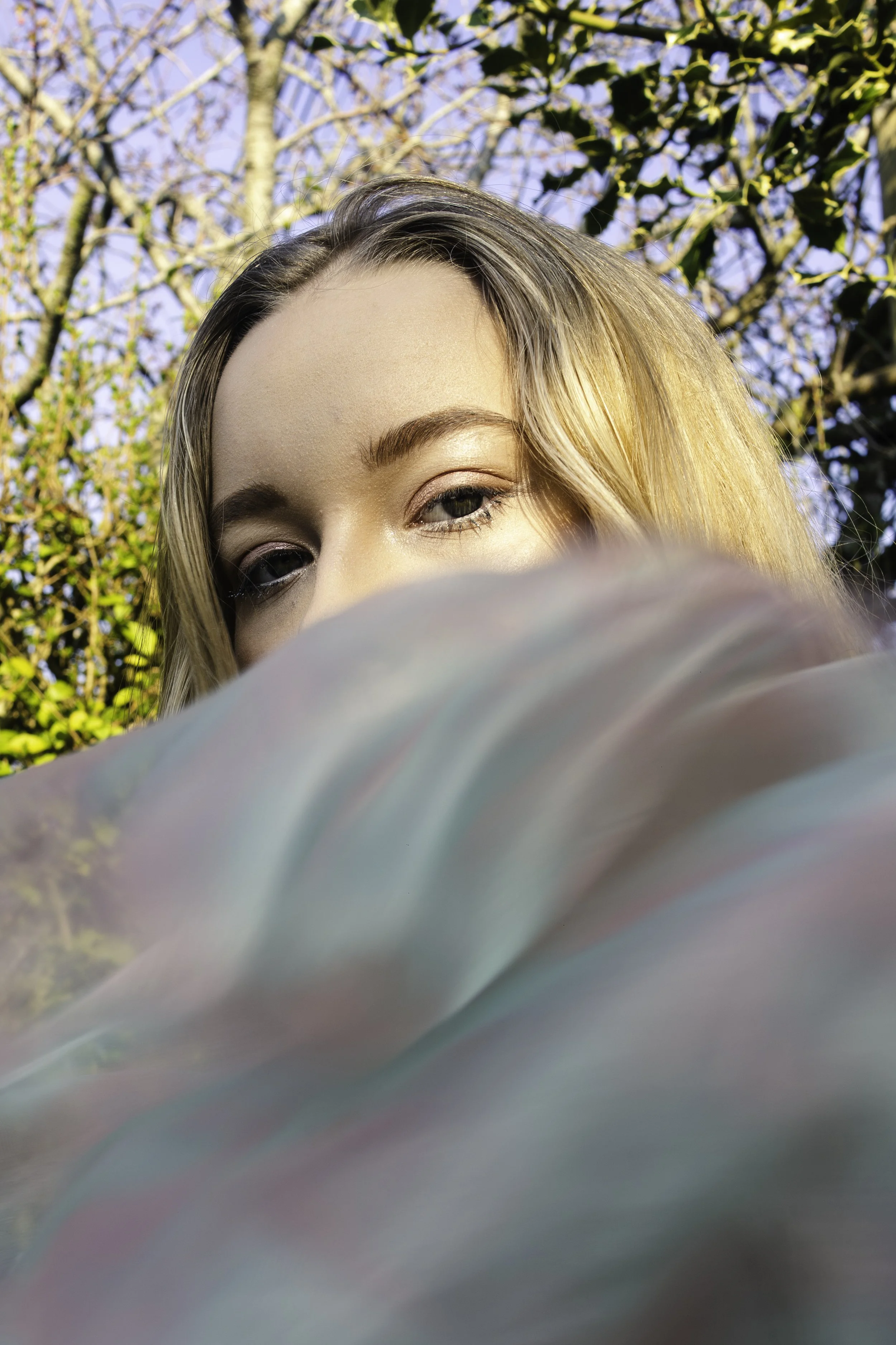 Close-up of a woman's face partially obscured, with her eyes and forehead visible, outdoors with trees in the background and sunlight on her hair.