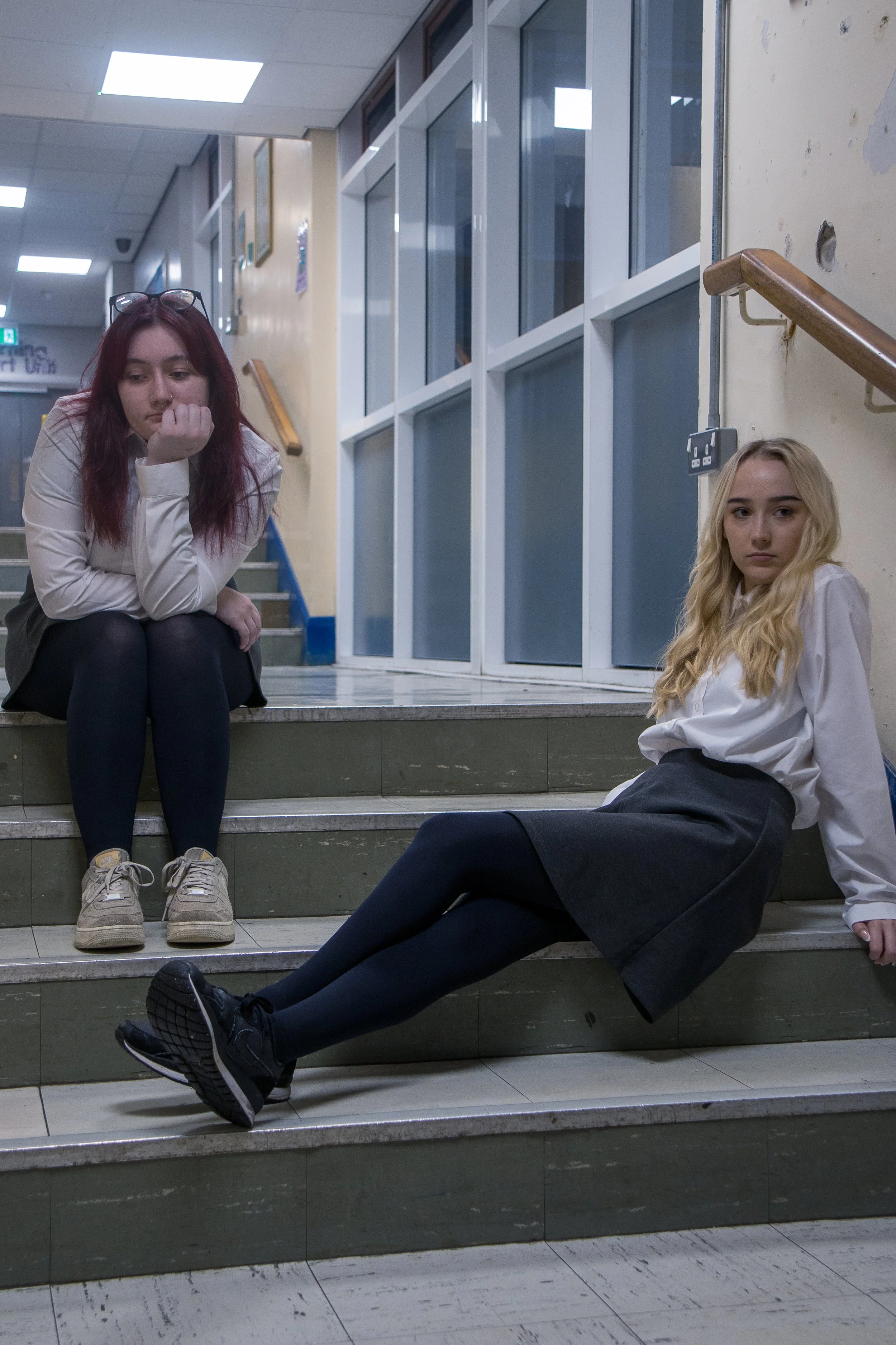 Two young women in school uniforms sitting on stairs in a school hallway. The woman on the left has long red hair, glasses on her head, and is resting her chin on her hand. The woman on the right has long blonde hair and is sitting on the stairs with