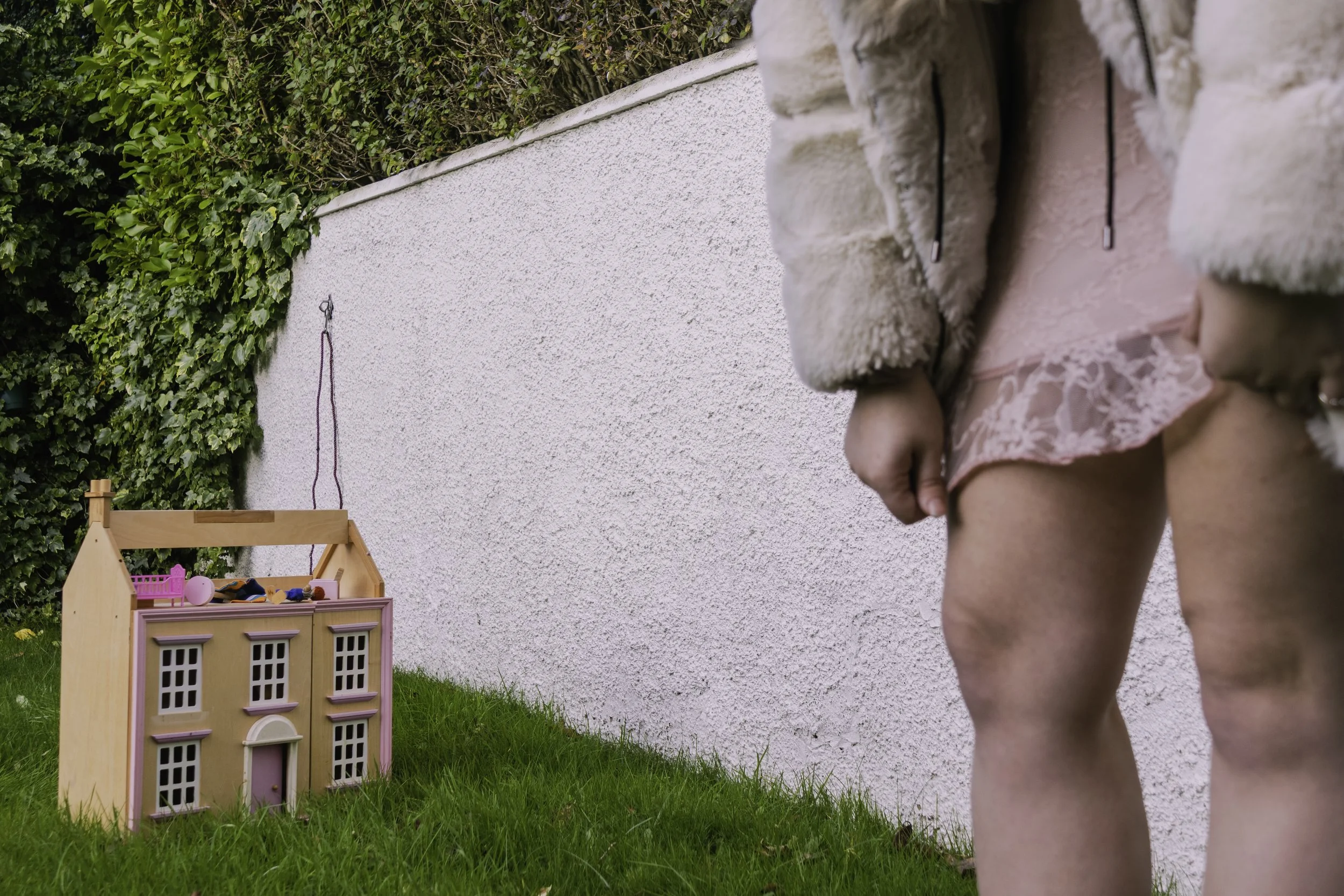 Woman playing outdoors next to a dollhouse with green grass and hedge in the background, wearing a pink dress and furry coat.