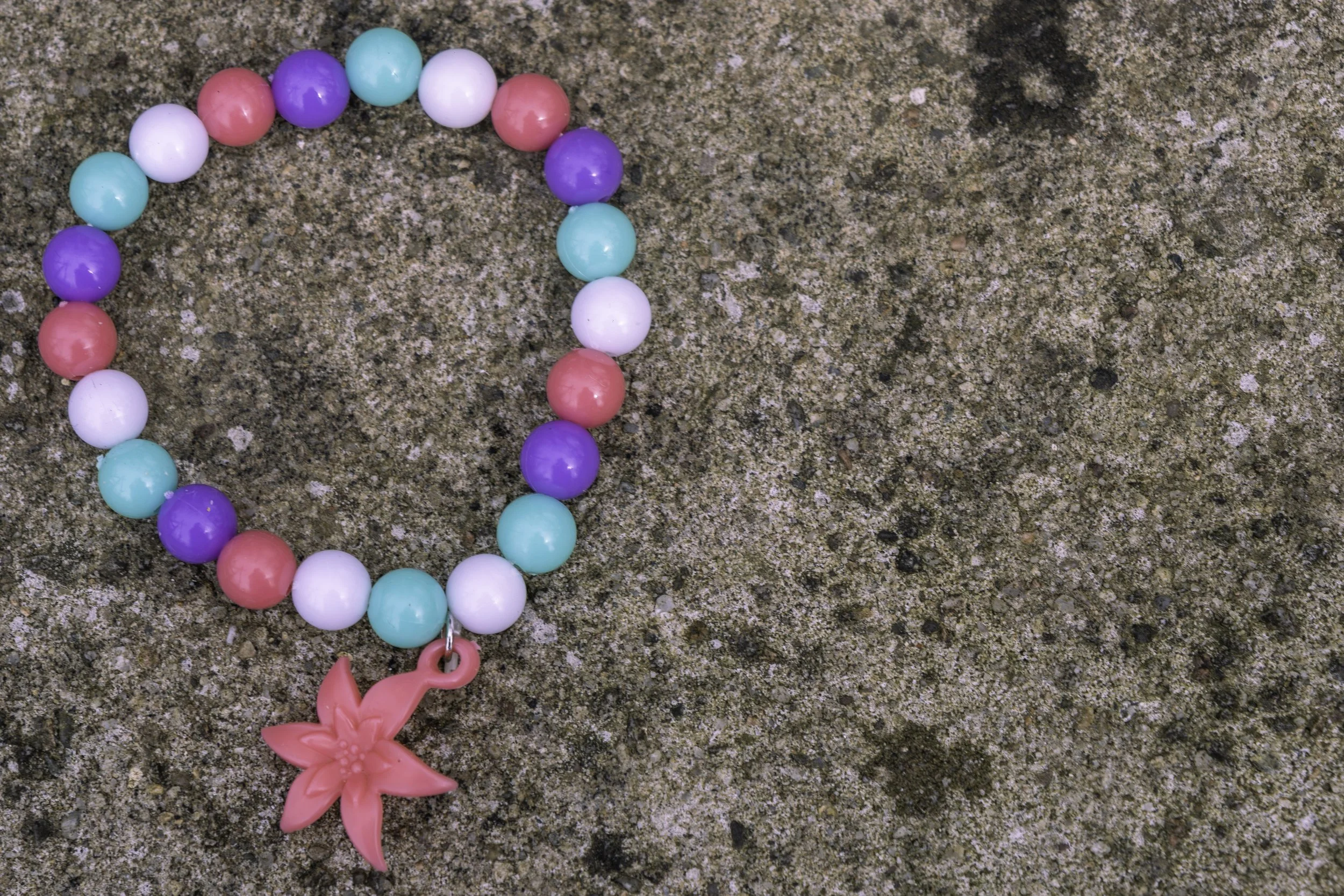 Colorful beaded necklace with a pink flower pendant on a sandy surface.
