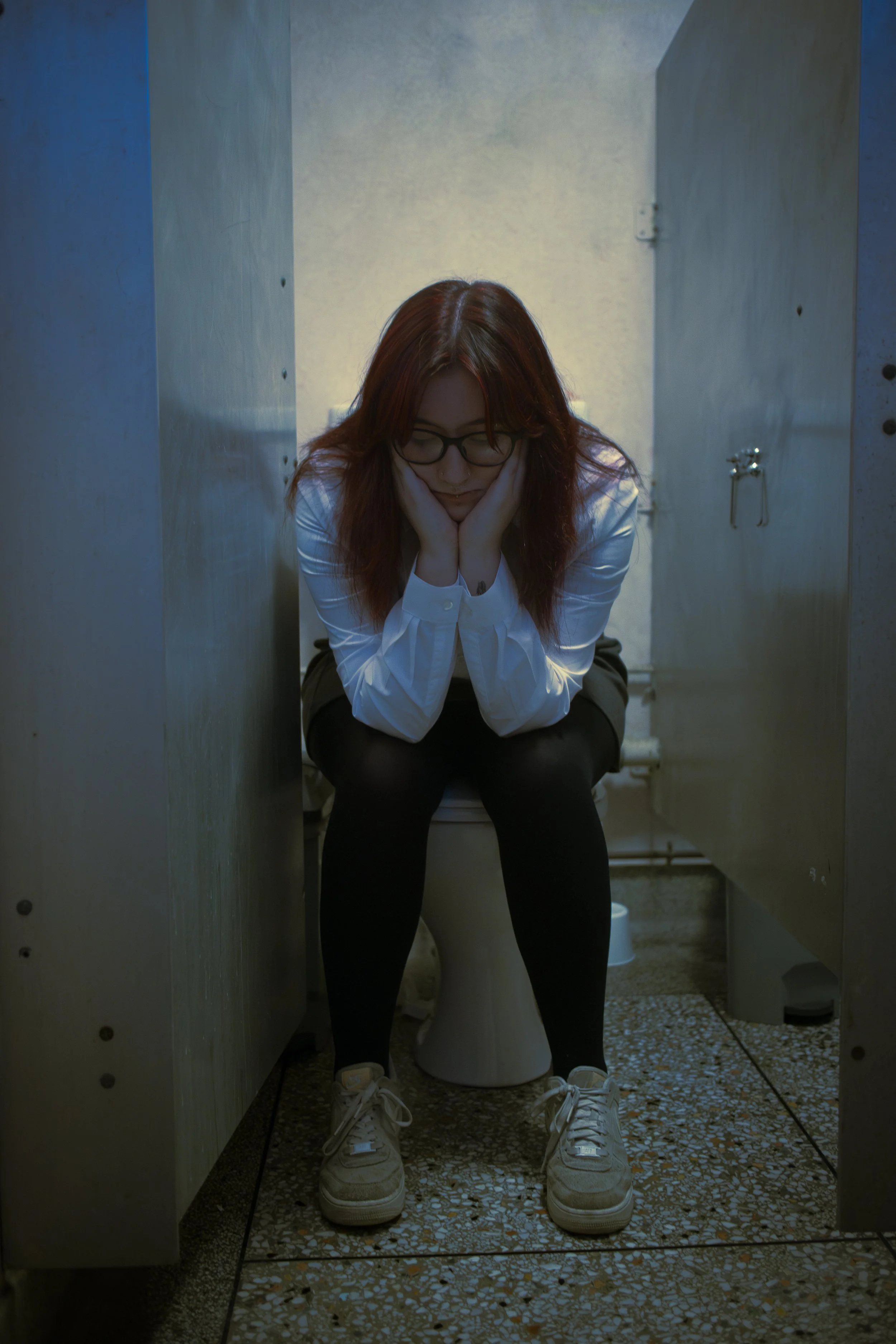 A woman with red hair and glasses sitting on a toilet in a bathroom, with her hands on her cheeks and kneeling with her feet flat on the ground.