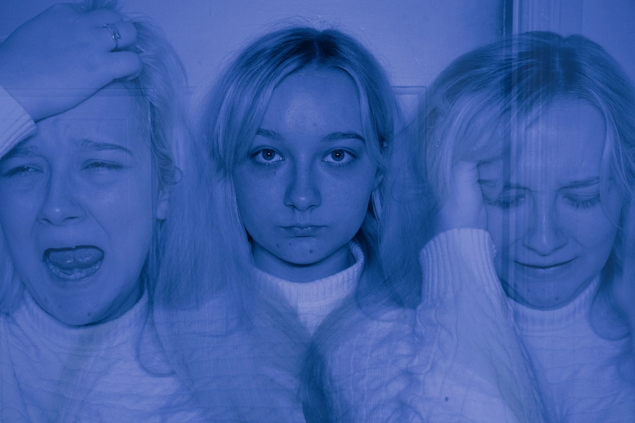 A person with long blond hair looking directly at the camera with a neutral expression, flanked by two masks with exaggerated facial expressions, all under blue lighting.