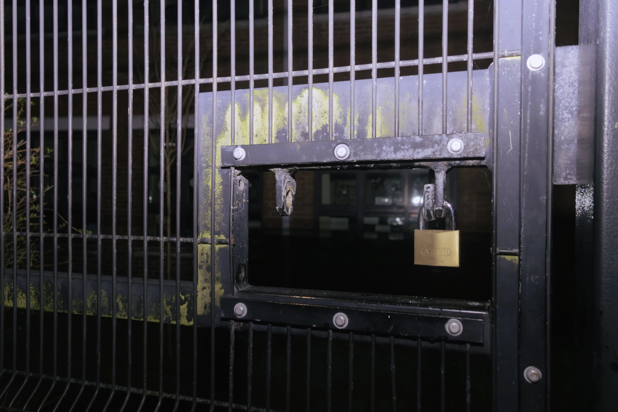 Close-up image of a black metal gate with a lock and a yellowly stained sign attached to it, with a brick building and windows visible in the background.