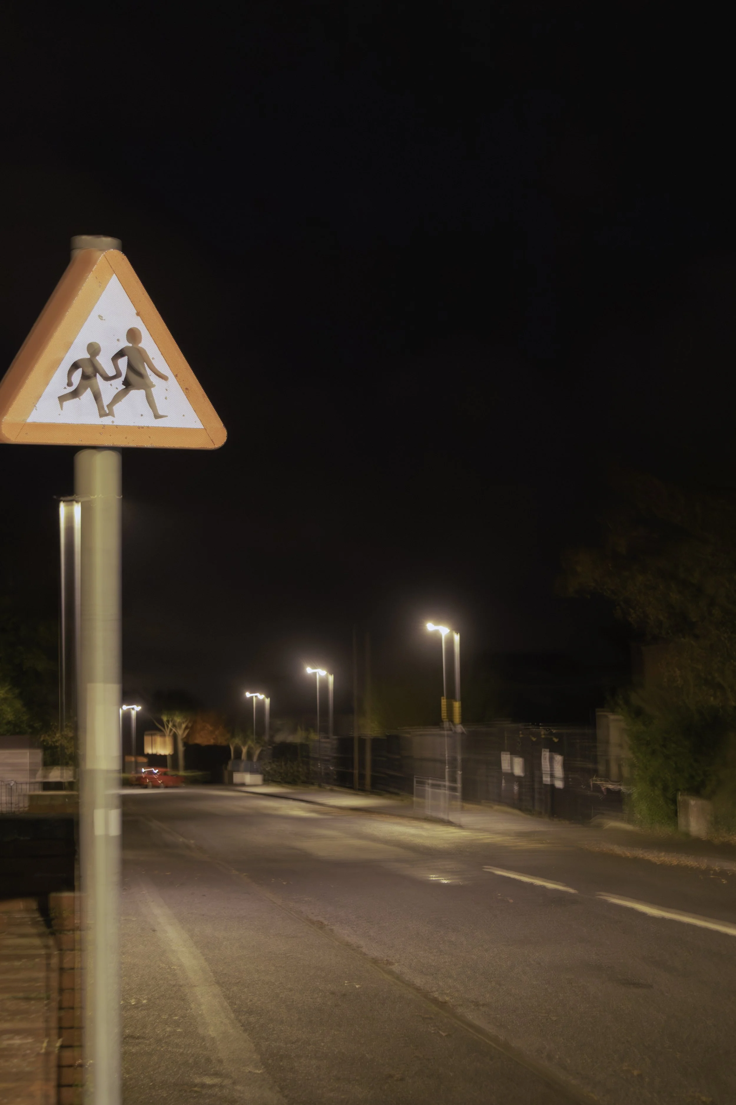Nighttime street scene with a crosswalk sign showing two children holding hands, illuminated by streetlights along the empty road.