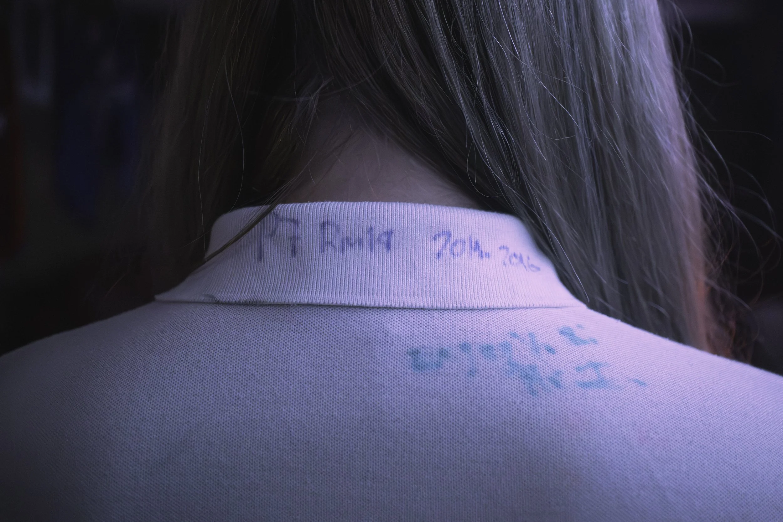 Close-up photo of the back of someone's head with long, straight hair, wearing a white shirt with handwritten notes in blue ink on the collar area.