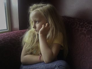 A young girl with long blonde hair sitting by a window, gazing outside with a thoughtful expression.