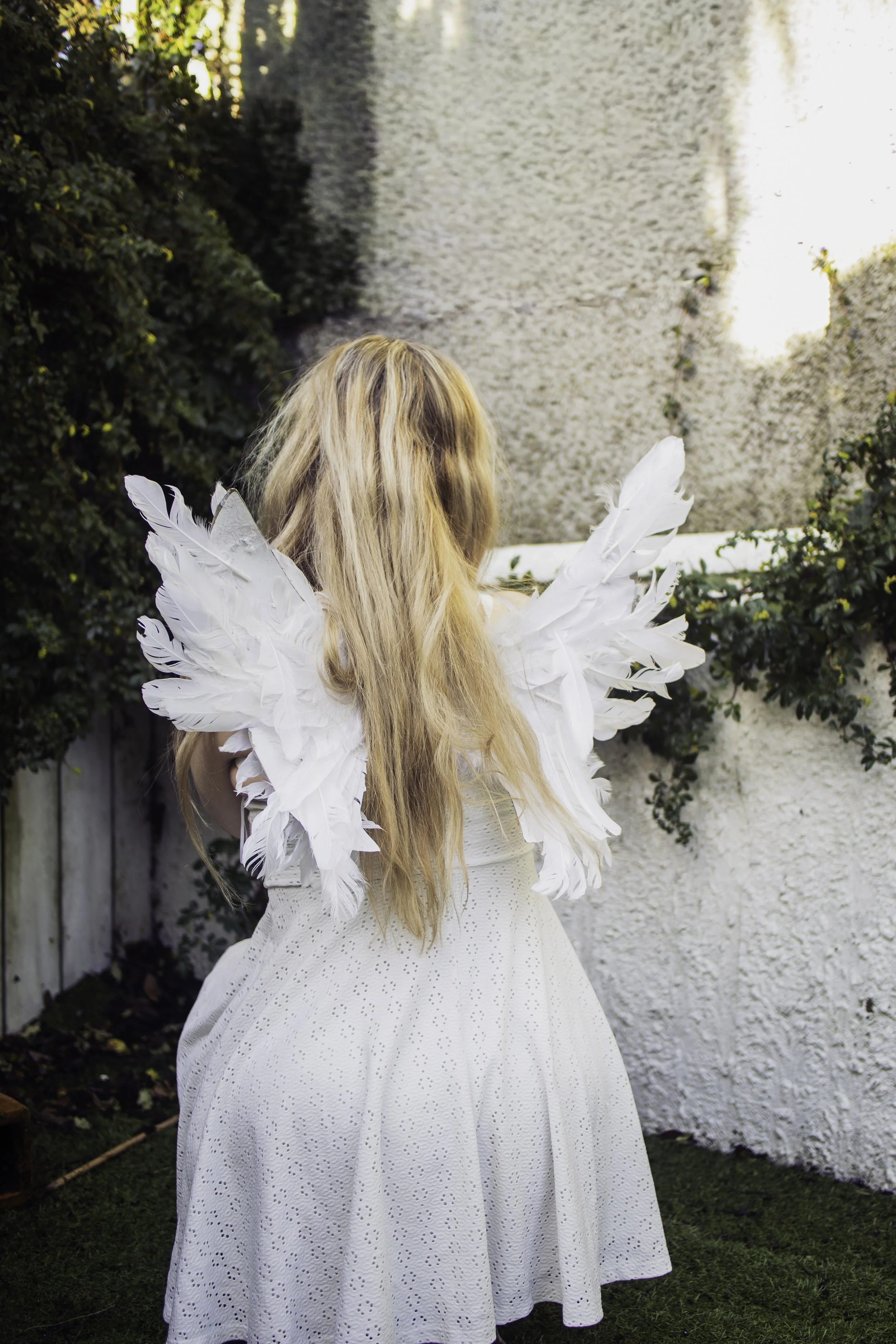 A person with long blonde hair dressed as an angel in a white dress with white feathered wings standing outdoors near a white wall with greenery.