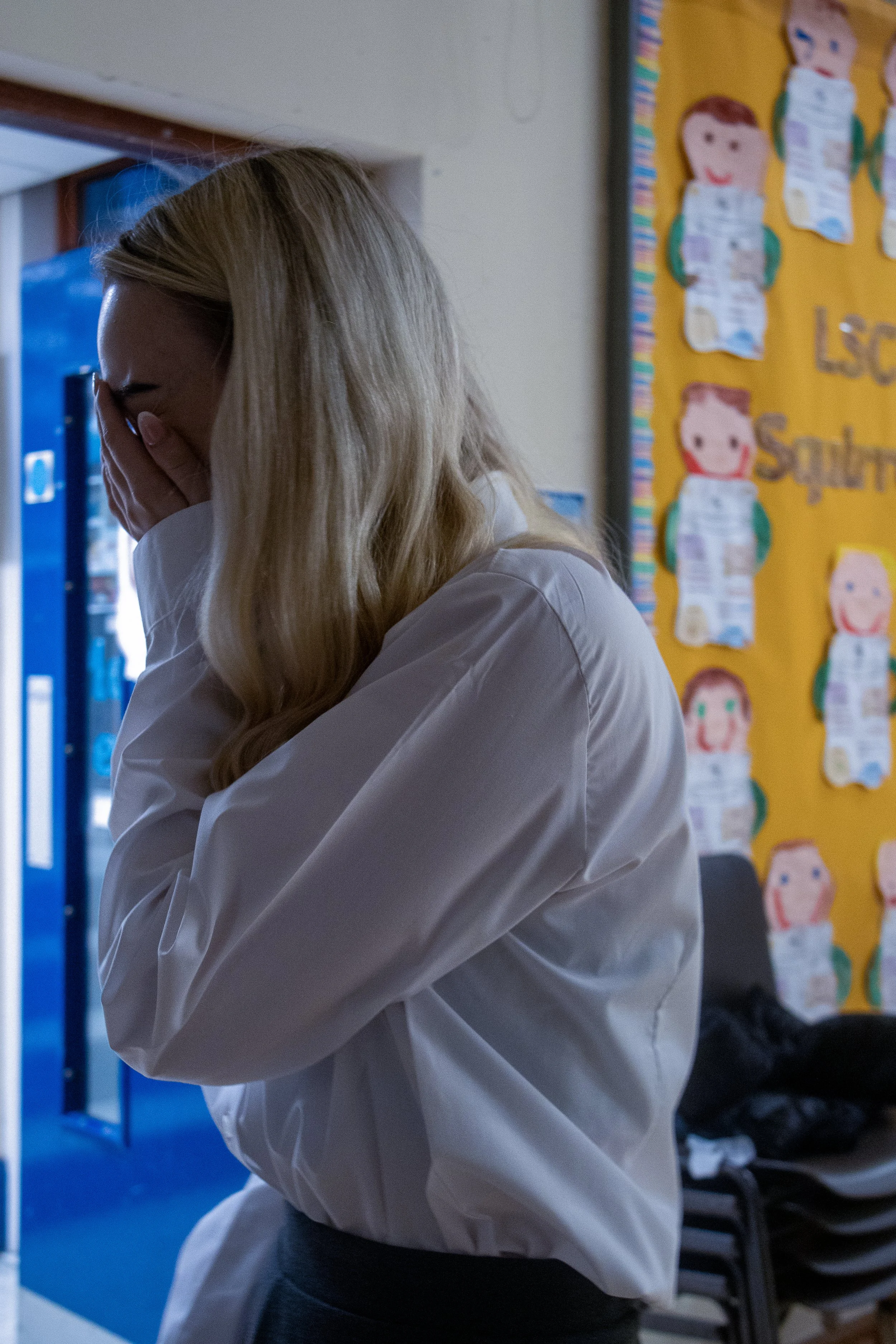 A woman with long blond hair wearing a white long-sleeve shirt and gray pants, standing indoors with her face covered by her hand.